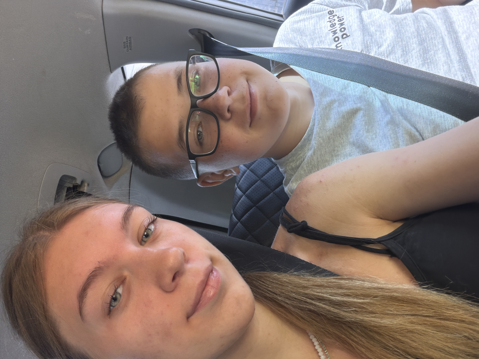 Two children, a girl and a boy, are seated in the front passenger seats of a car. The girl, on the left, has blonde hair and is wearing a black tank top with thin straps and a pearl necklace. The boy, on the right, is wearing glasses and a light gray t-shirt with the text "Knowledge Power" printed on it. Both are wearing seatbelts. The interior of the car, including the seats and headrests, is visible. The scene is likely taking place during the daytime, as indicated by the natural lighting. The image does not provide any specific details that would allow for the location to be identified as Rostov-on-Don, Russia.