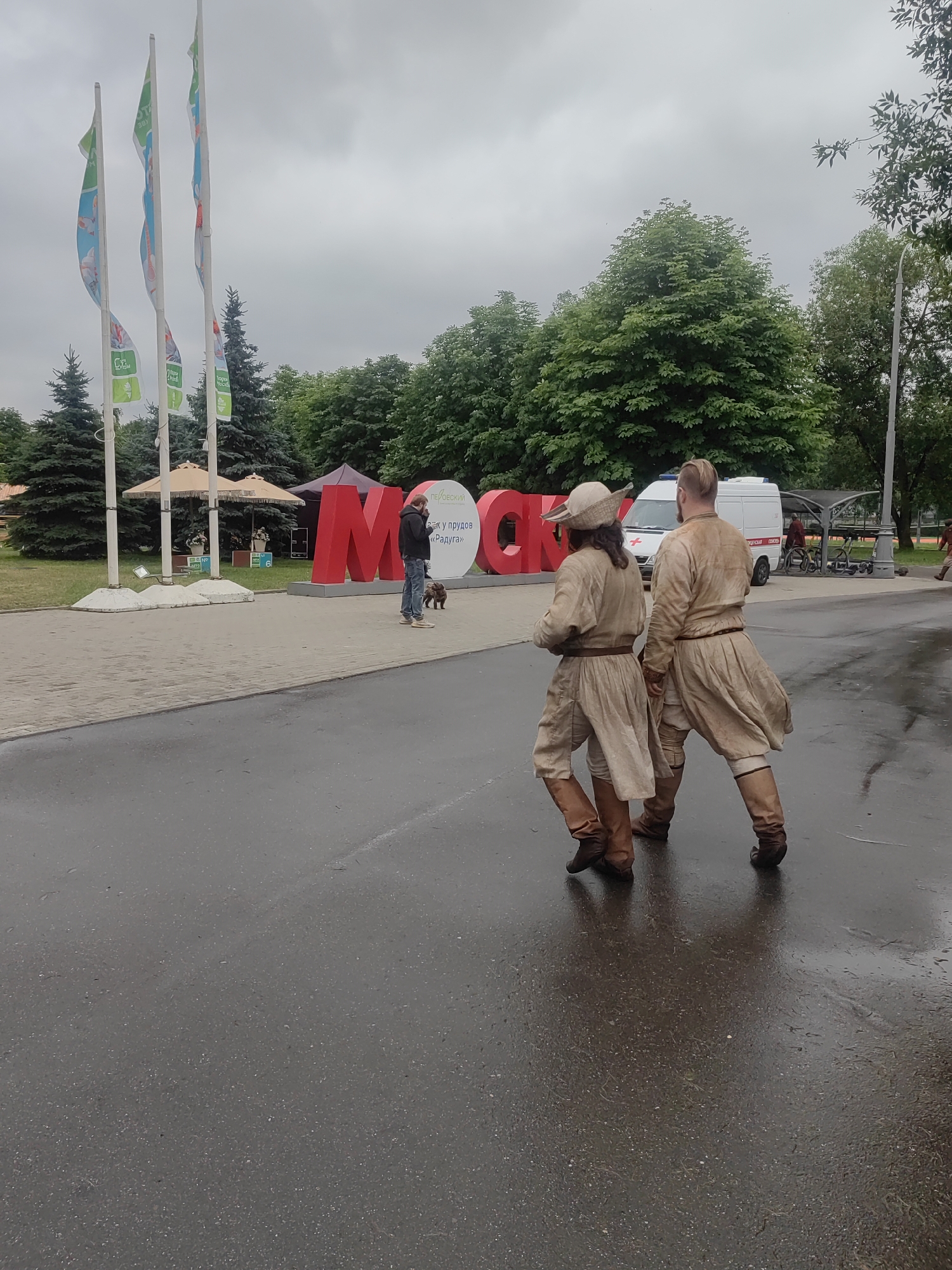 Two individuals in historical reenactment attire, one wearing a hat, walk on a wet paved surface. The man on the right has a visible top knot hairstyle. Behind them, large red letters spelling "MOSCOW" are visible, with smaller text in Russian on the 'W'. In the background, several white flags on tall poles are erected, and a white van with a red cross emblem is parked. Green trees form a backdrop, and the sky is overcast. A person with a dog is also visible near the large letters. The scene appears to be an outdoor event or park in Moscow, Russia.