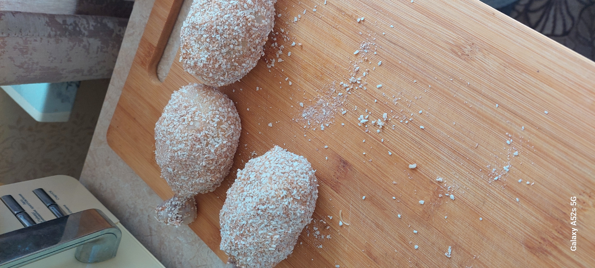 Three food items, appearing to be raw chicken legs coated in breadcrumbs or a similar coating, are arranged on a wooden cutting board. The cutting board is positioned on a countertop, and a portion of a kitchen appliance is visible in the upper left corner of the image. Scattered breadcrumbs are present on the cutting board around the food items.