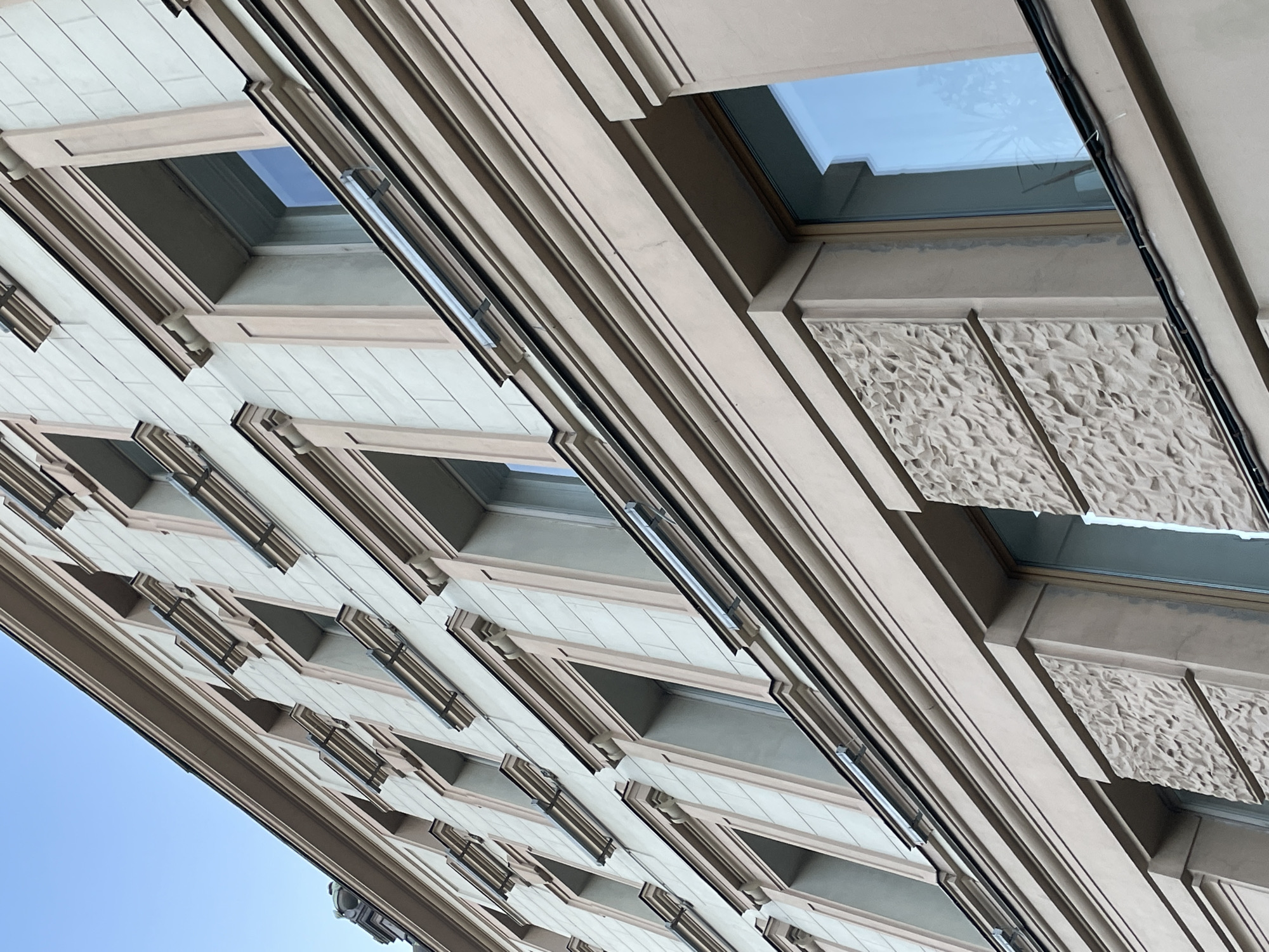 The image shows the exterior facade of a building, likely located in Saint Petersburg, Russia. The building has multiple windows, arranged in a repeating pattern. The windows are rectangular with dark frames, and the facade is decorated with textured panels between the windows. The photograph is taken from a low angle, looking up at the building. The sky is visible in the lower left corner. A few cables are running along the right side of the building.