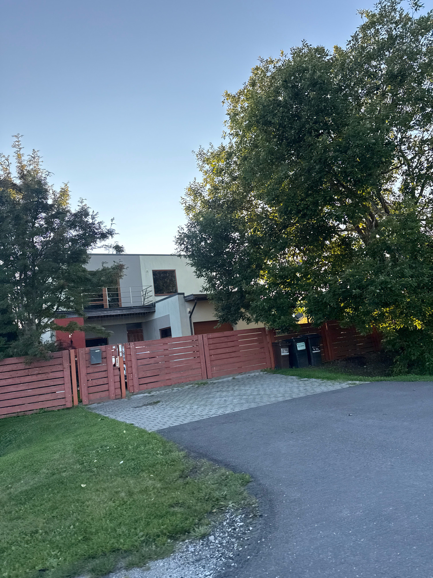 The image shows a modern, two-story house with a light-colored facade and dark-framed windows, located in Peetri alevik, Estonia. A red wooden fence encloses the property. To the left of the house, there is a tall evergreen tree, and to the right, a large deciduous tree with lush green foliage. The area in front of the fence is paved with light gray interlocking stones, and a patch of green grass is visible to the left of this area. A paved road runs in the foreground, curving towards the right side of the image. Three dark-colored trash bins are placed near the fence on the right side. The sky is clear and blue, suggesting it is daytime. The scene appears to be a residential street.