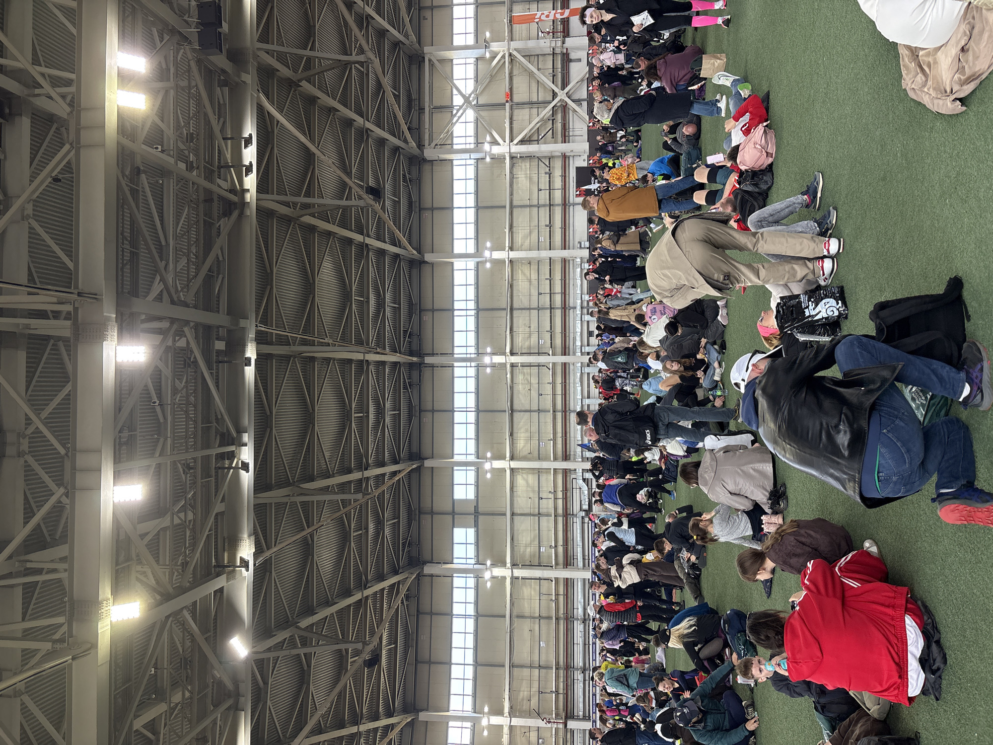 The image shows a large group of people seated on a green surface in a vast indoor space. The space appears to be an indoor arena or gymnasium, suggested by the visible roof support structure with metal beams and industrial lighting. The people are densely packed together and facing in the same general direction. Many of them are looking down, possibly at something in their laps. Clothing varies among individuals. Based on the crowd size and indoor sports flooring, it is plausible that this photo was taken at the Метеор stadium, Матюшино, Russia.