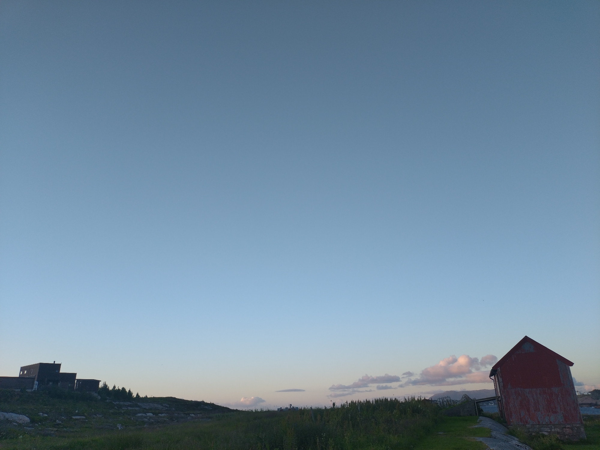 The image shows a rural landscape at dusk in Averøy, Norway. On the left, a dark, multi-segmented building complex is perched on a rocky, grassy incline, with a line of dark green trees behind it. On the right, a tall, red, corrugated metal barn stands at an angle. The sky is a pale blue with scattered, soft pink and white clouds on the horizon, suggesting the sun has recently set or is about to rise. The foreground consists of green grass and rocky terrain. The scene is devoid of people or any visible activity.