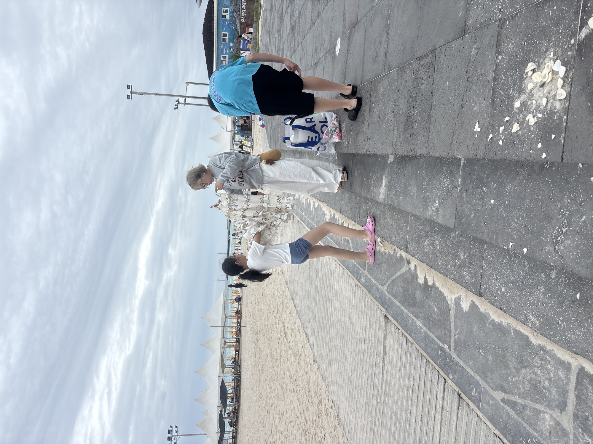 The image depicts a scene on a cloudy day at a beach in Jeju, South Korea. In the foreground, on a paved walkway, a young girl in a white t-shirt and blue shorts stands facing away from the camera, holding a white garment. To her right, an adult wearing a grey sweatshirt and white pants examines the same garment. To the right of the adult, a person in a bright blue t-shirt and black shorts bends over, appearing to interact with the ground. A large, white reusable shopping bag with blue lettering is visible between the adult and the person bending over. In the background, a sandy beach is lined with white tent-like structures and lounge chairs. Further back, a building with blue accents and a mural is visible, along with power lines and wind turbines. The ground in the foreground is paved with large, dark gray stones and there are scattered white shells on the pavement.