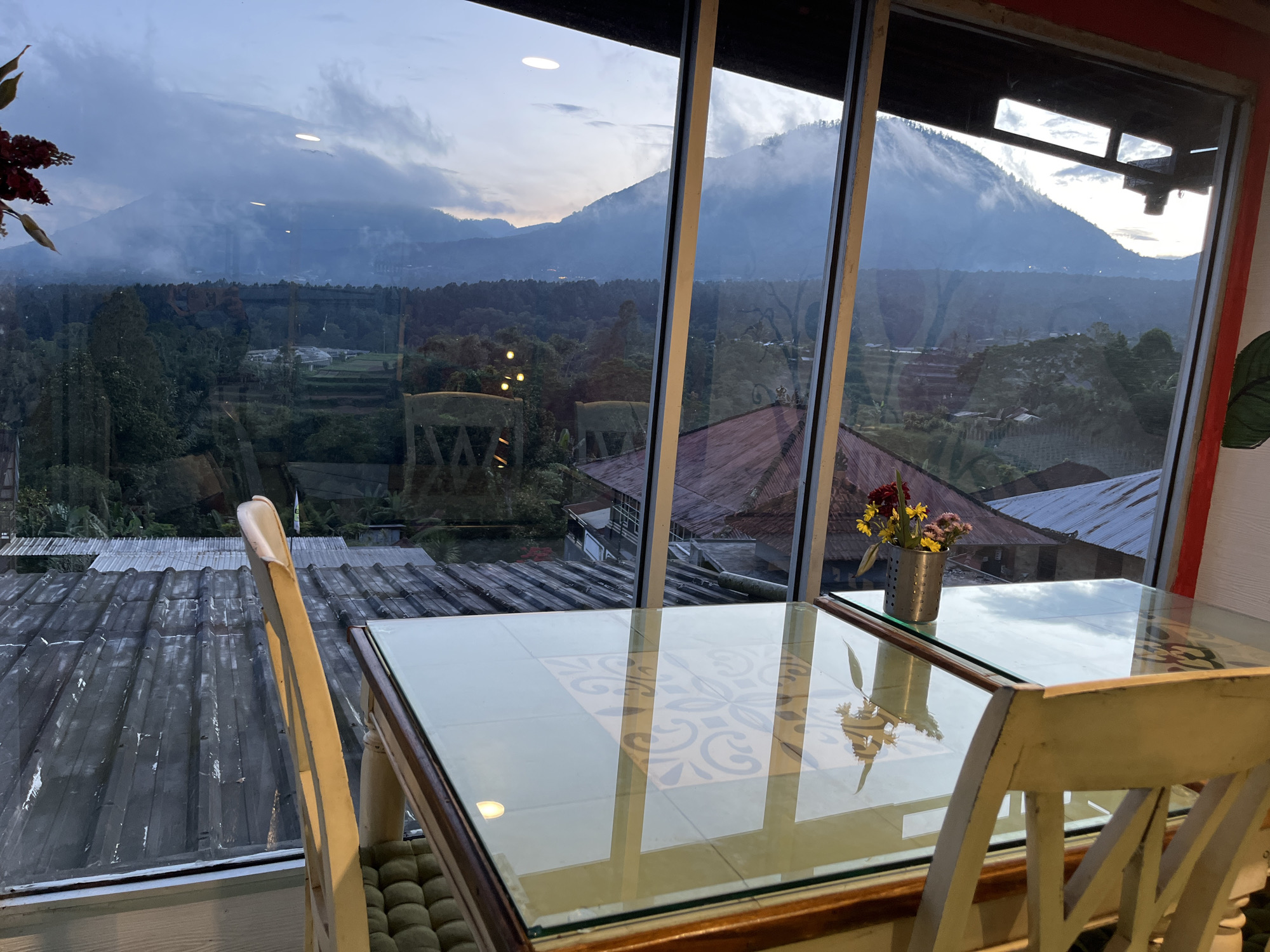 A daytime indoor scene shows a window view of Baturiti, Indonesia. Visible through the window is a mountainous landscape under a cloudy sky, with trees and buildings in the foreground. Inside, a glass-topped table with a floral design is surrounded by two light-colored wooden chairs. A small vase with flowers sits on the table. A metal roof is partially visible below the window.