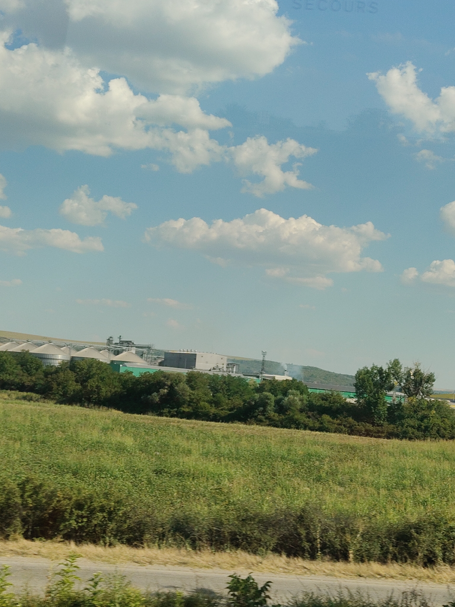 The image captures a broad landscape scene under a partly cloudy sky, with the text "SECOURS" faintly visible in the upper portion of the sky. In the mid-ground, an industrial complex is situated behind a line of lush green trees. The complex features several large, conical silos, a grey rectangular building, and other structures. To the right of the industrial area, a tall green structure is visible, partially obscured by trees. Further to the right, a solitary green tree stands out. In the foreground, a vast field of green vegetation extends towards the viewer, with a grey road running horizontally across the bottom of the frame. The location is identified as Yasen, Bulgaria.