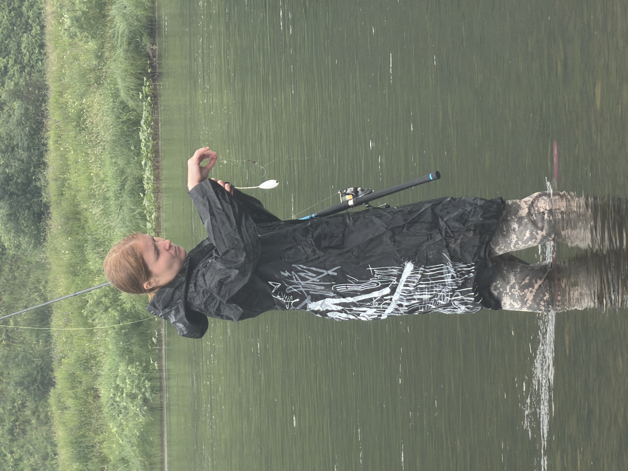 A person stands in the shallow water of a river, likely in Ust-Kabirza, Russia, facing right and holding a fishing rod. The person is wearing a black raincoat with white graffiti-style markings and camouflage waders. They are holding a fishing lure and line in their right hand, examining it. The river water is calm and green, with a lush, grassy bank visible in the background. The sky is overcast.