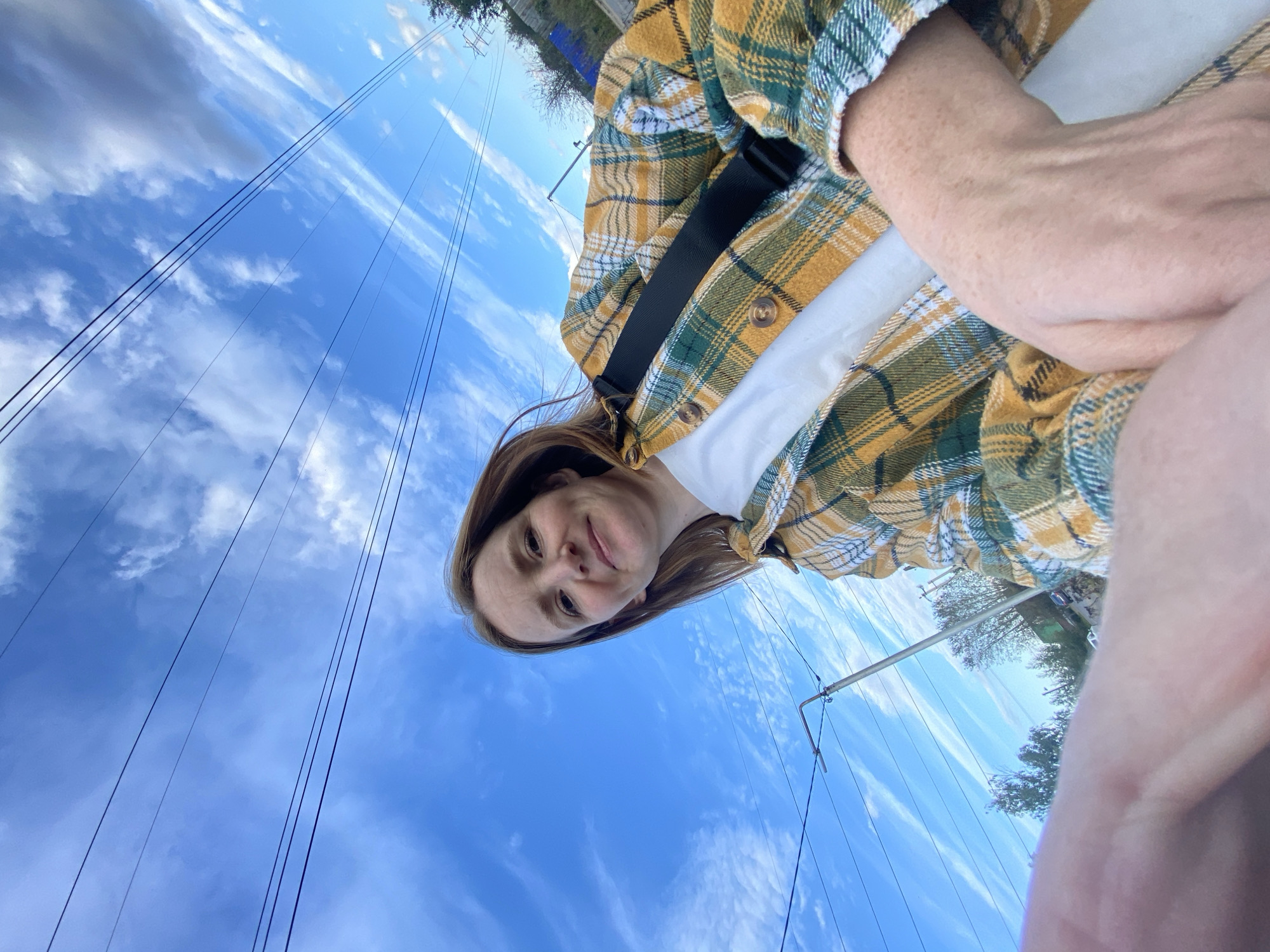 A low-angle selfie shows a woman with auburn hair and fair skin, freckled cheeks, and a slight smile. She is wearing a white t-shirt and an open yellow and green plaid shirt. A black strap is visible across her chest. Her left hand is extended towards the camera, partially obscuring the bottom left corner. The background consists of a bright blue sky with wispy white clouds, intersected by several power lines running diagonally. In the distance, there are trees, a utility pole, and a fence, suggesting an outdoor urban or semi-urban setting. Based on visual cues such as architecture and vegetation, and with additional information from external sources, this image is likely located in Chaikovsky, Russia.