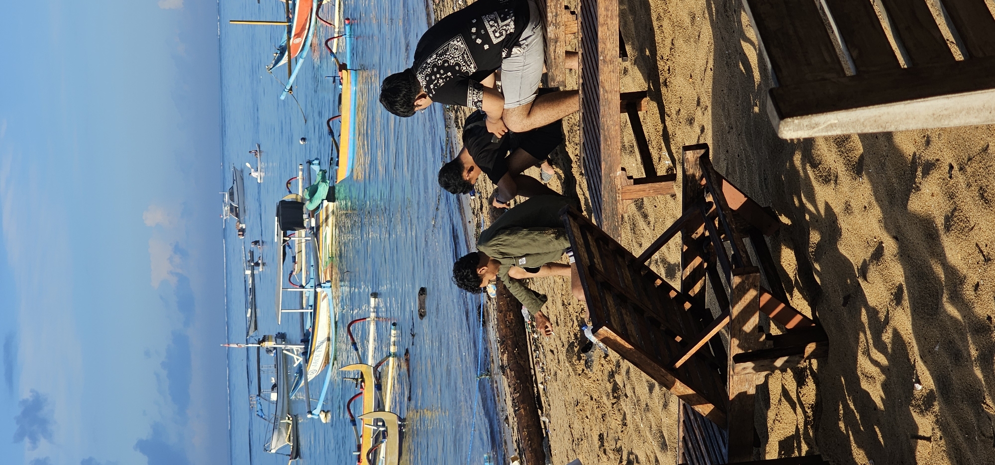 Three young men are on a sandy beach in Denpasar, Indonesia. They are at the water's edge, near wooden boats anchored in the shallow, blue water. The man closest to the viewer wears an olive-green shirt, the next wears a black shirt, and the third wears a black shirt with a white design and gray shorts. A wooden structure with a table and steps is on the sand near the men. The sky is blue with scattered clouds.