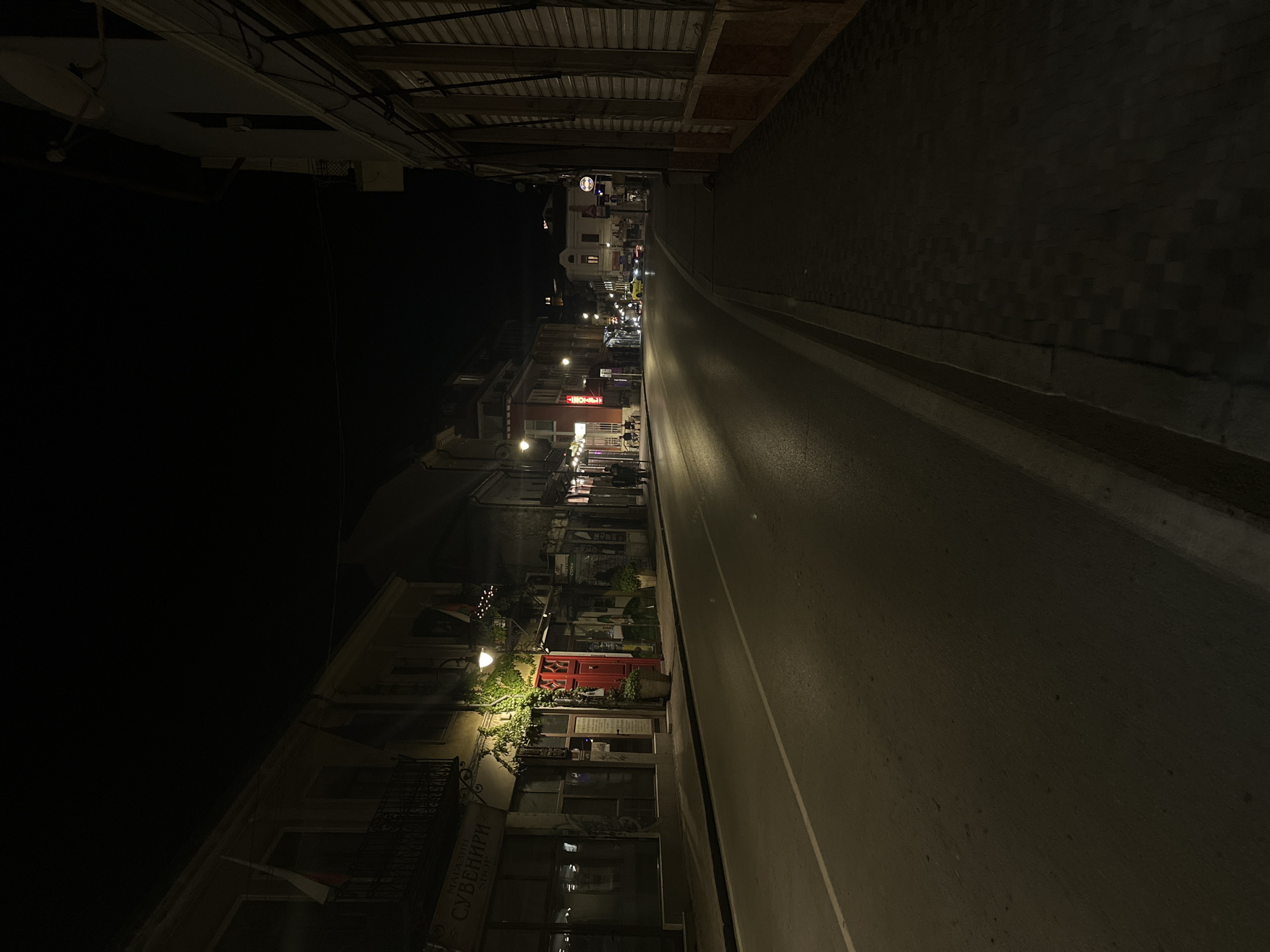 The image shows a nighttime view of a street in Veliko Tarnovo, Bulgaria. The street is illuminated by streetlights and light emanating from storefronts. Buildings line the left side of the street, with shops and restaurants visible. A red door can be seen on one of the buildings. The right side of the street appears to be a stone wall. The street slopes upward into the distance. The scene is dark, with only the street and buildings lit.
