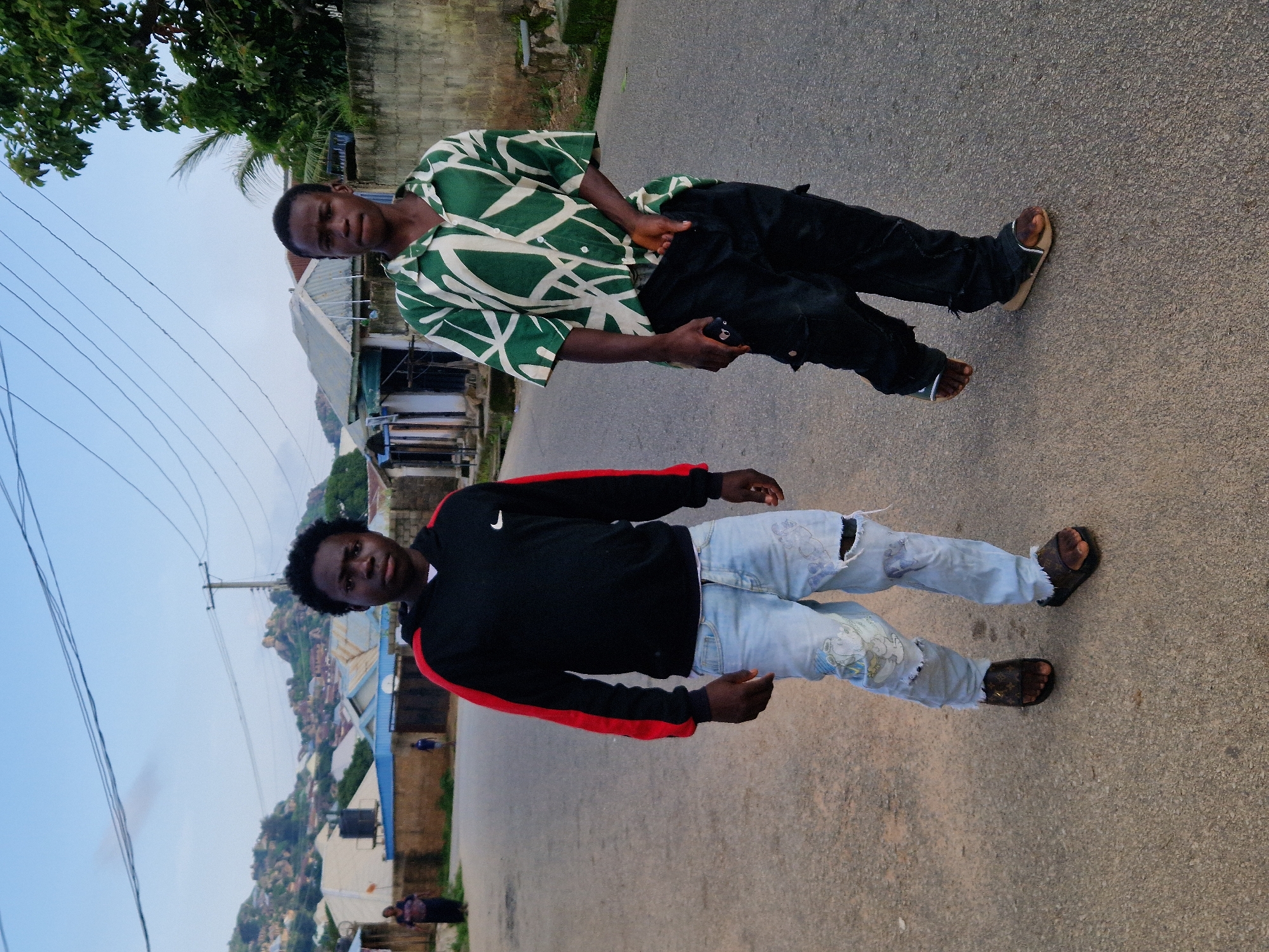 Two young men walk on an asphalt road in the city of Kabong, Nigeria. The man on the left wears a black hoodie with red sleeves and light blue ripped jeans with cartoon graphics. He has dark skin and short black hair and is wearing brown sandals. The man on the right wears a short-sleeved green and white patterned shirt and black cargo pants. He has dark skin and short black hair and is wearing brown sandals. The background shows a street with houses and trees. The sky is partly cloudy.