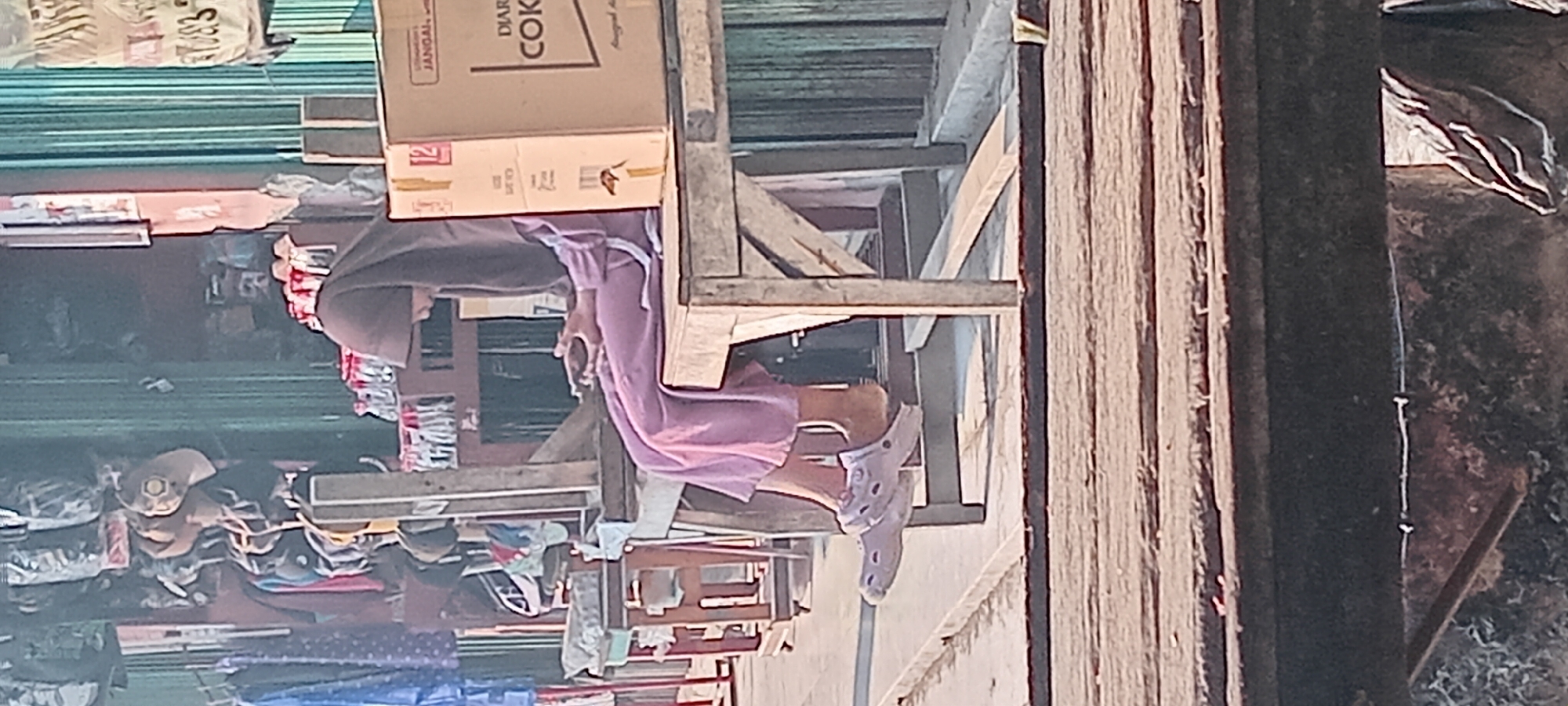 A side-view, medium shot shows a person wearing a hijab and a light purple dress sitting on a wooden bench. The person is holding a mobile phone in their hands. The person is wearing light purple Crocs-style sandals. The background is blurred but suggests an outdoor market with stalls displaying various items like clothing, bags, and boxes. One visible box has the word "DIAR" and "COK" printed on it, suggesting it contains a beverage. The setting appears to be the city of Karangsari, Indonesia.
