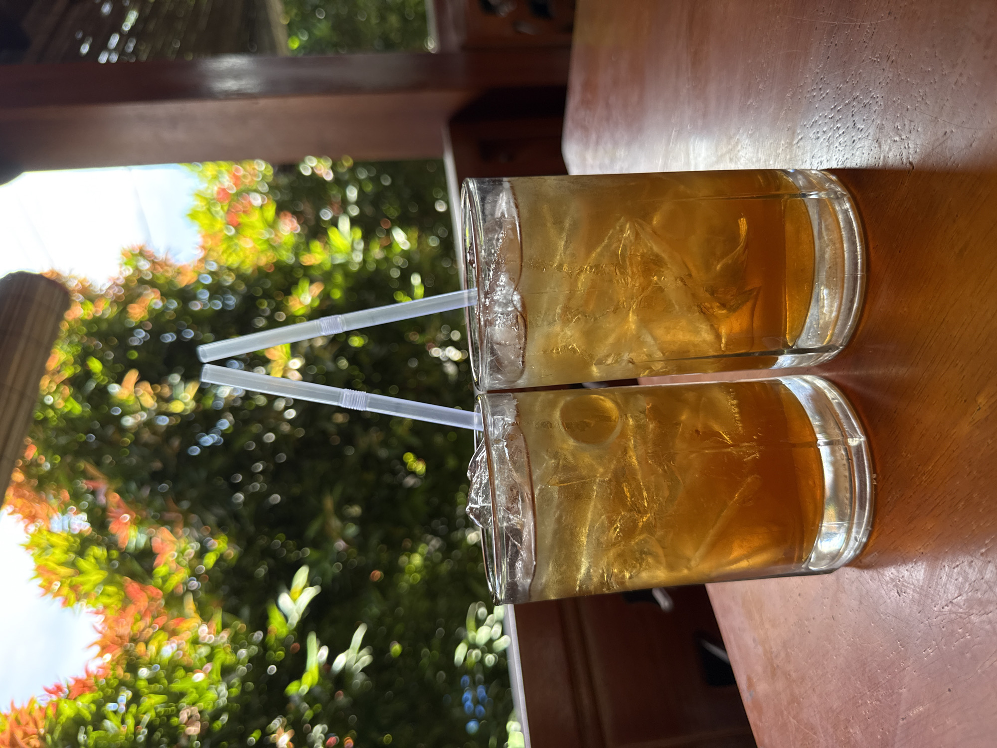 Two glasses filled with ice and a yellow liquid sit on a wooden table. Each glass has a straw. The background shows a blurry view of trees with green and red leaves, suggesting an outdoor setting. The image is taken from a close perspective, focusing on the drinks and the immediate surroundings. The location is Lhokseumawe, Indonesia.