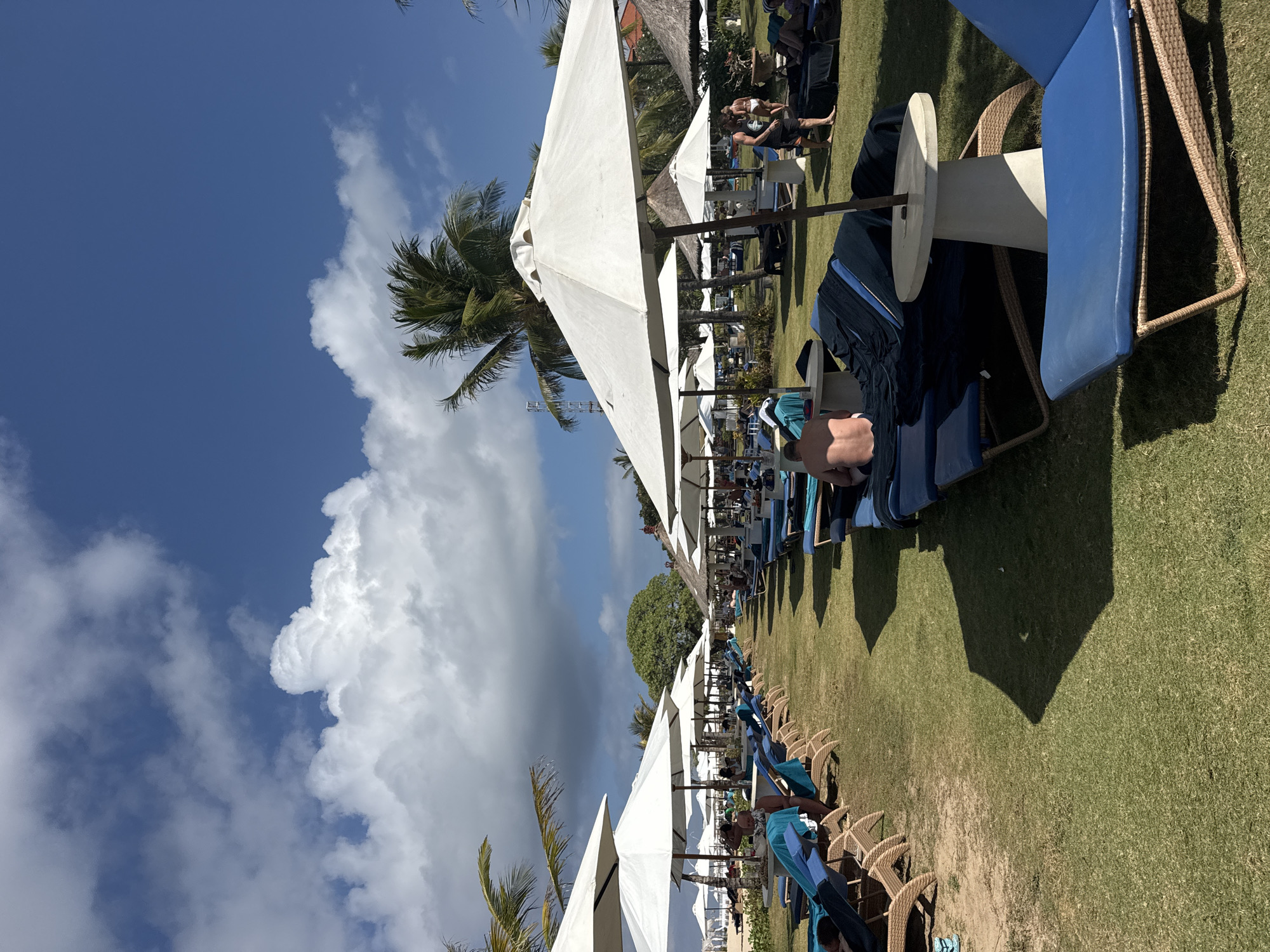 This image depicts a well-maintained resort area in Kuta Selatan, Indonesia, characterized by rows of blue lounge chairs covered with towels, arranged on a lush green lawn. White umbrellas are strategically placed to provide shade for the loungers. In the background, palm trees and other tropical foliage suggest a warm climate. Several individuals are visible, some relaxing on the chairs, others standing and conversing, and a few are seen walking. The sky is a vibrant blue with scattered white clouds, indicating fair weather. The scene conveys a sense of leisure and relaxation typical of a holiday destination.