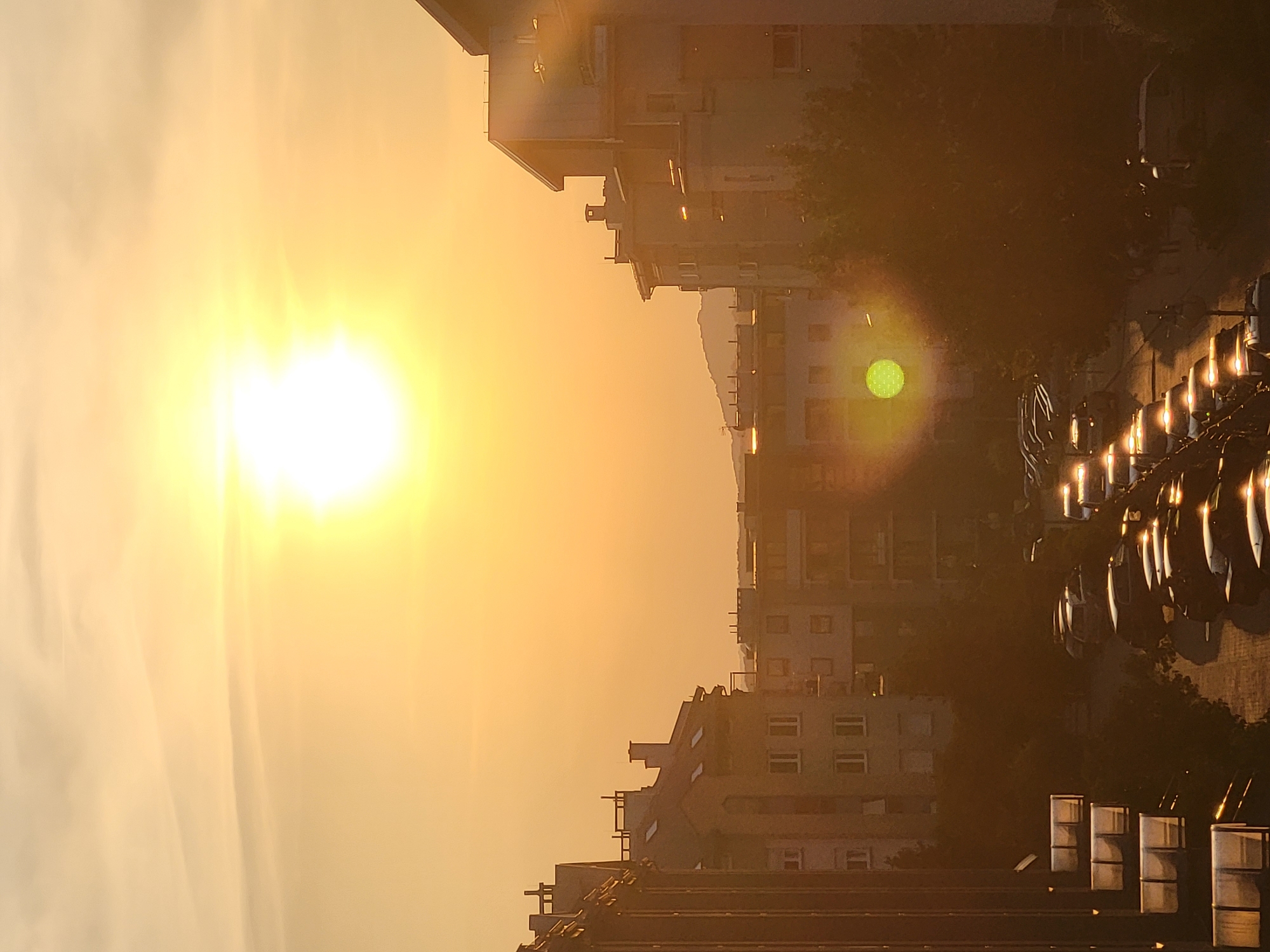 The image depicts an urban landscape at sunset, with the sun appearing as a bright, diffused orb in the sky, casting a warm orange glow. The scene is dominated by apartment buildings, visible on both the left and right sides of the frame, as well as in the background. The architecture suggests a residential area, with multiple windows and balconies on the buildings. In the foreground, a street is lined with parked cars, their headlights illuminating the road. Trees are scattered throughout the scene, adding a touch of nature to the urban environment. The overall impression is one of a peaceful evening in a city.