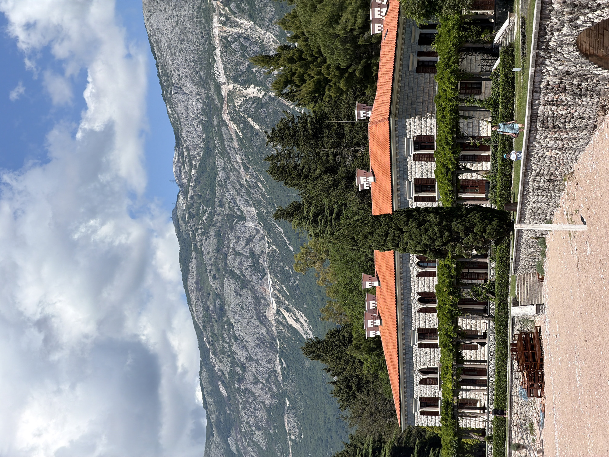 The image presents a view in Sveti Stefan, Montenegro, focusing on structures positioned against the backdrop of a mountain range and partly cloudy sky. Two light-colored buildings with orange tile roofs stand parallel to each other, featuring evenly spaced windows with brown shutters. Lush green vines grow along the building facades. Adjacent to the buildings is a rocky pathway, where two people can be seen. One person is wearing a blue dress and the other is wearing a blue cap. The surrounding environment includes dense green trees and vegetation separating the buildings from the mountain. The sky overhead is partly covered with clouds.