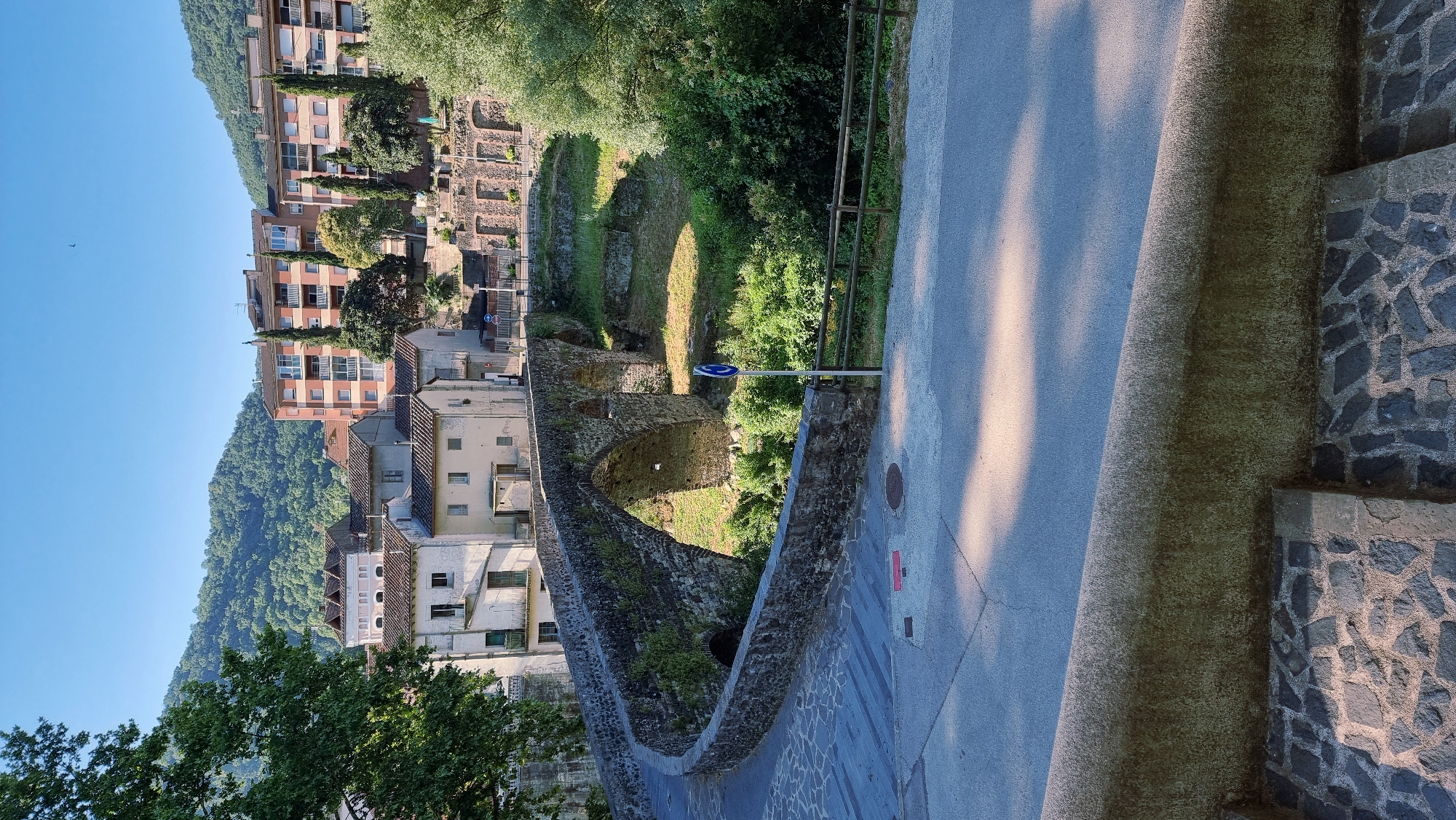 This is a photograph taken in the city of Sant Joan les Fonts, Spain, capturing an ancient stone bridge arching over a dry riverbed. The bridge is made of large, uneven stones, some of which are covered in moss, indicating age and exposure to the elements. The bridge has a curved approach on the left side, paved with cobblestones, which transitions to a modern paved road in the foreground.
Behind the bridge, a cluster of buildings is visible. These include older, light-colored houses with tiled roofs, and a taller, more modern apartment building with balconies. The architecture suggests a blend of historical and contemporary styles. The surrounding landscape is hilly and covered in dense green vegetation, characteristic of the Garrotxa region. The sky is clear and blue, indicating fair weather.
In the riverbed, patches of dry grass and scattered greenery are visible. A metal fence runs along the bank of the riverbed, separating it from the road. A blue sign with a white arrow indicating a directional turn is planted near the bridge. The foreground shows a paved area with shadows cast by trees or structures, suggesting it is daytime. The lower part of the image displays a stone wall and a set of stone steps. The overall scene depicts a scenic view of a historic landmark integrated into a residential and natural environment.