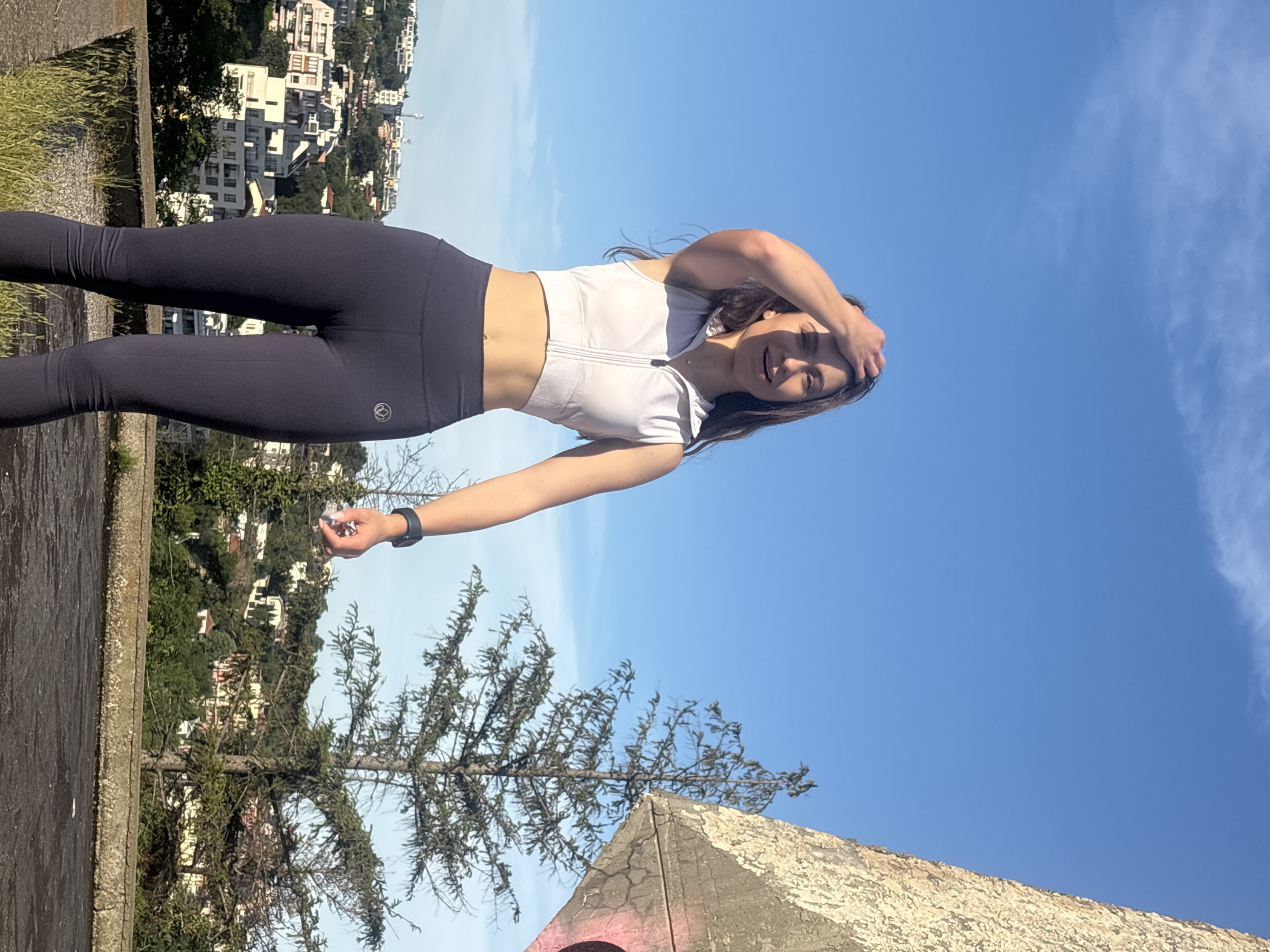 A young woman with long, dark hair stands outside with her hand in her hair, smiling at the camera. She is wearing a white, sleeveless zip-up crop top and dark leggings. She has a watch on her left wrist. Behind her, the sky is blue with a few scattered clouds. In the background, a city with buildings and trees is visible on a hill. To the right of the woman, a large, abstract concrete structure is partially visible, with a tall evergreen tree in front of it. The scene appears to be a city overlook in Varna, Bulgaria.
