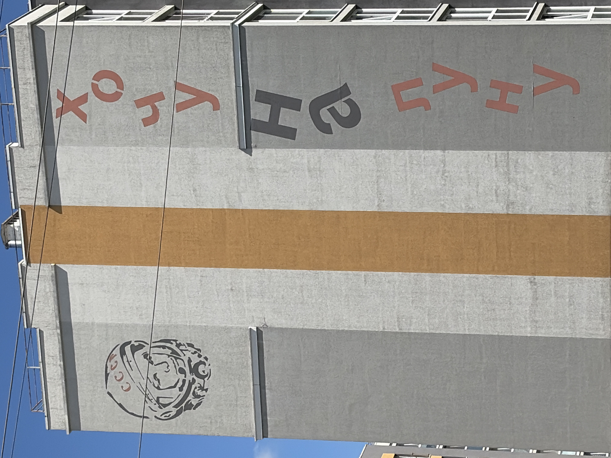 This media file captures a close-up, eye-level view of a tall, grey building facade against a clear blue sky. The building appears to be a modern structure with a flat roofline featuring horizontal window elements.
Dominating the upper portion of the facade are large, stylized letters painted in red and dark grey. On the left, in red, are the characters "x o y". To their right, in dark grey, are the characters "H a". Further to the right, also in red, are the characters "L y H y". The letters are bold and geometric in design.
A wide, horizontal band of warm brown color runs across the middle of the building, dividing the upper section with the letters from the lower section. Below this band, the building's surface is a lighter shade of grey.
In the lower left quadrant of the image, a circular emblem is painted in black and red. The emblem depicts a stylized face within a complex, gear-like or helmet-like design. Red lettering, possibly "CCCP," is visible within the emblem's border.
The presence of electrical wires running vertically in front of the building suggests an urban or semi-urban environment. The bright sunlight and clear sky indicate it is daytime with fair weather. The overall impression is one of public art or signage on the exterior of a building.