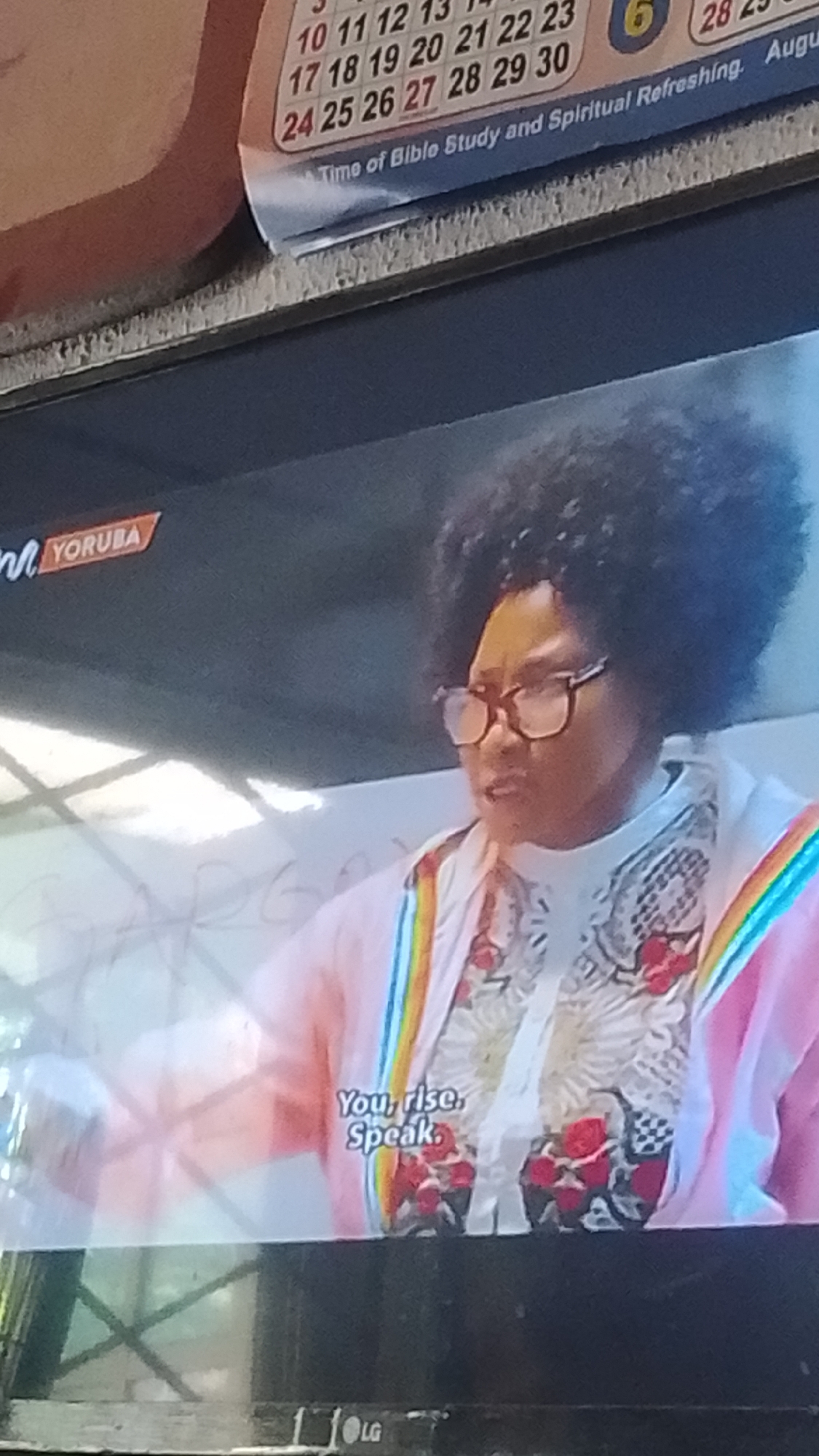 A television screen displays a close-up shot of a woman with a large afro hairstyle, wearing glasses and a brightly colored jacket. The jacket has rainbow stripes on the collar and cuffs. Beneath the jacket, she wears a patterned shirt with floral designs. Text on the screen reads "YORUBA" in the upper left corner, and "You, rise. Speak." in the lower right. A calendar with dates and the inscription "Time of Bible Study and Spiritual Refreshing. August" is visible at the top of the screen, suggesting a potential educational or religious context. The LG logo is visible at the bottom of the screen. The scene appears to be indoors, possibly a studio or a room with a whiteboard in the background, though the background is largely out of focus. The lighting is bright, creating a somewhat harsh contrast. The woman's expression is serious as she looks slightly off-camera. The image quality is grainy, typical of a photograph taken of a screen. There is no visible information that suggests the location is specifically Lokoja, Nigeria.