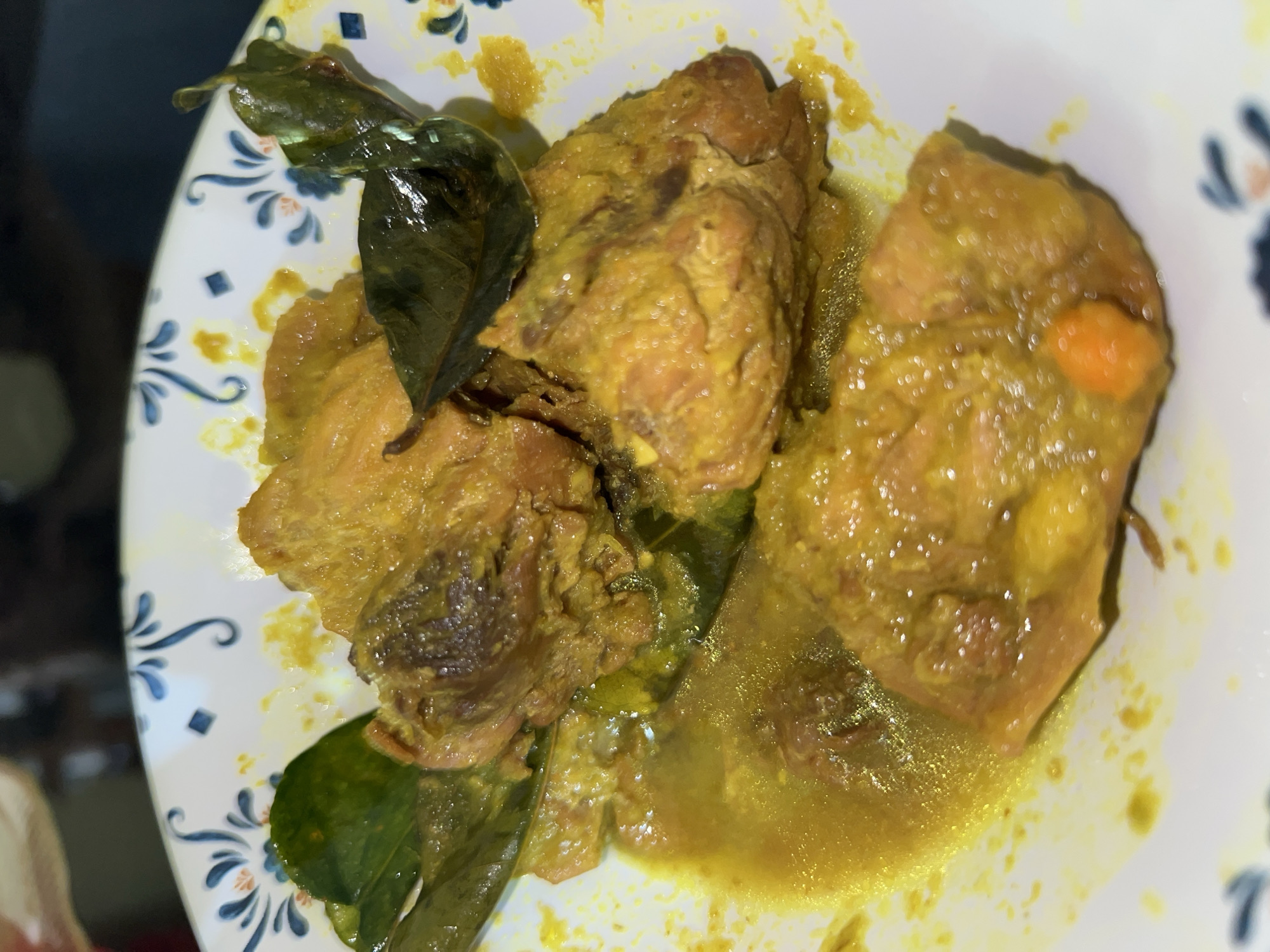 A close-up, overhead shot shows a white plate with a blue and white floral pattern containing a serving of chicken curry. The chicken pieces are coated in a thick, yellow curry sauce and appear tender. Several dark green leaves, possibly kaffir lime leaves, are interspersed with the chicken. The plate is filled with the curry, which has a glossy sheen. A small, orange piece, possibly a piece of chili or vegetable, is visible in the lower right portion of the dish. The background is dark and out of focus, suggesting an indoor setting. The location is specified as Klapanunggal, Indonesia.
