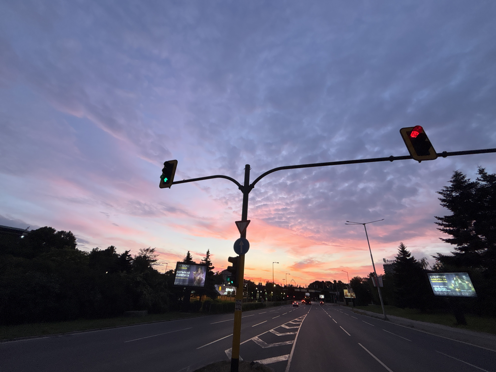 The image captures a view of a wide road in Sofia, Bulgaria, during a sunset. The sky is a dramatic canvas of purple, pink, and orange hues, with clouds streaking across. Traffic lights are visible, with one showing a red light and another a green light. A traffic sign with an upward arrow is also present. The road has multiple lanes and is lined with trees and streetlights. Billboards are also visible along the side of the road, with some displaying advertisements. A few cars are driving on the road. The overall scene suggests an urban environment experiencing the transition from day to night.