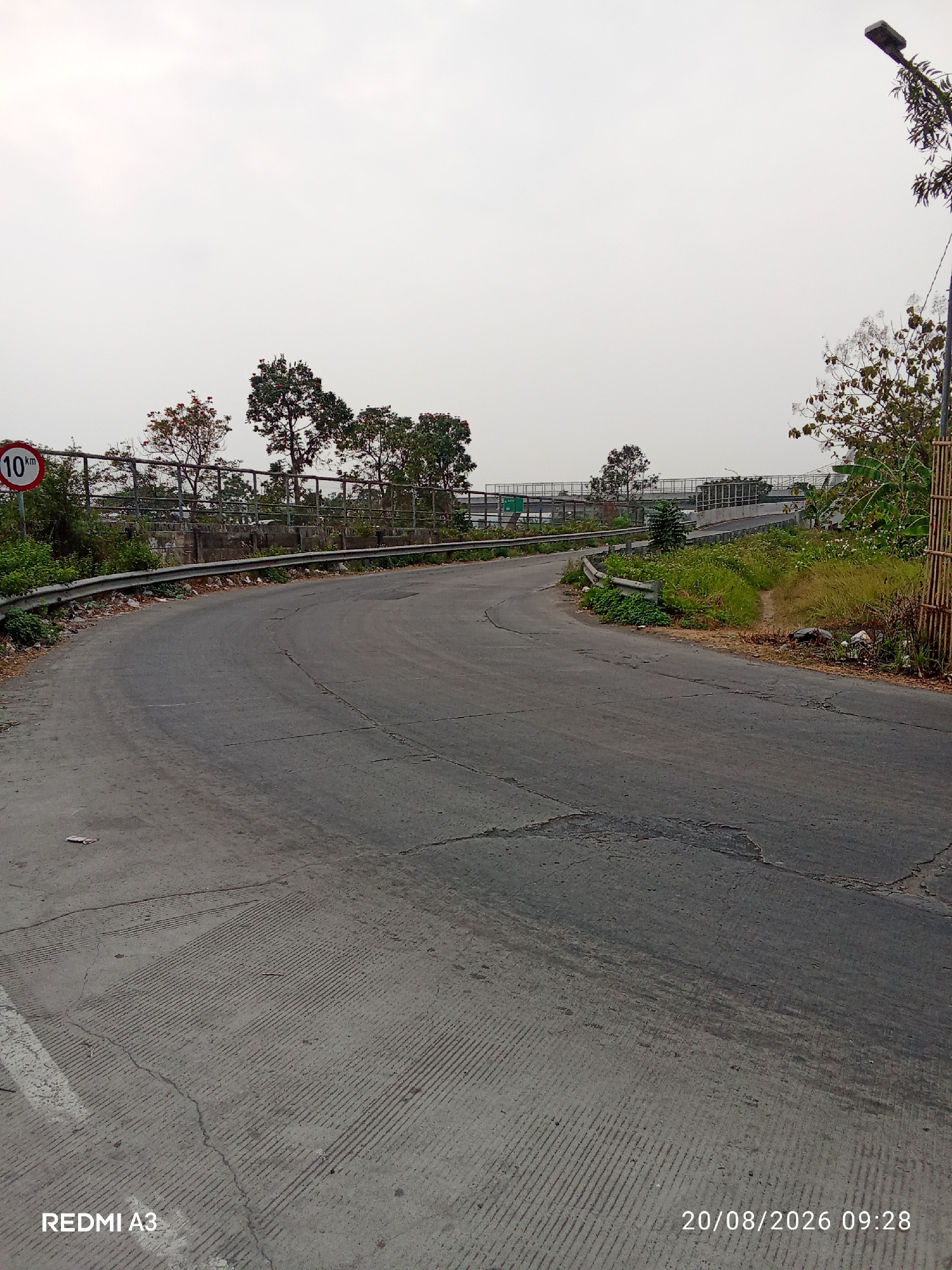 The image depicts a curved asphalt road in a semi-urban or rural setting, likely in Desa Pandeyan, Indonesia, given the timestamp. The road surface shows signs of wear, with cracks and patching. A speed limit sign indicating "10 km" is visible on the left side of the road, near a metal guardrail. Vegetation, including trees and bushes, lines the side of the road, with a concrete barrier and fence further back. In the distance, an elevated road or bridge structure is visible, suggesting a highway or overpass. A street light is present on the right side of the frame. The sky is overcast. The bottom of the image displays a watermark indicating "REDMI A3" and a timestamp of "20/08/2026 09:28". No people or specific actions are depicted in the scene.