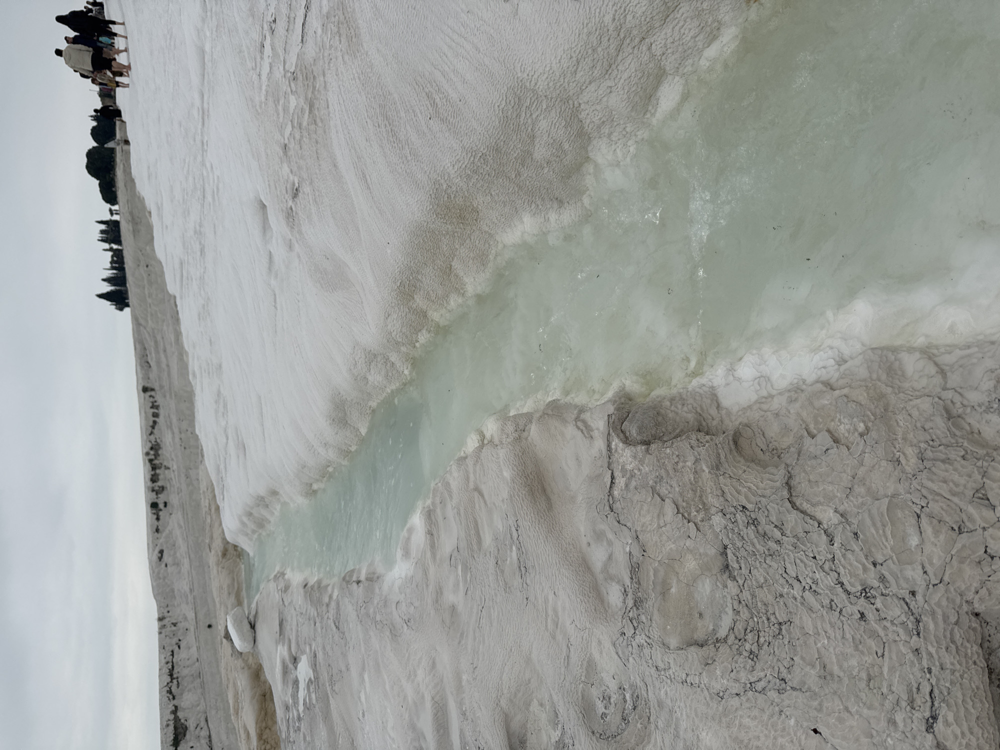 The image shows a section of the Pamukkale travertines in Turkey. The white, terraced formations are visible, with a shallow channel of turquoise water flowing through them. Some people can be seen in the distance, at the upper left side of the image. The day is overcast.