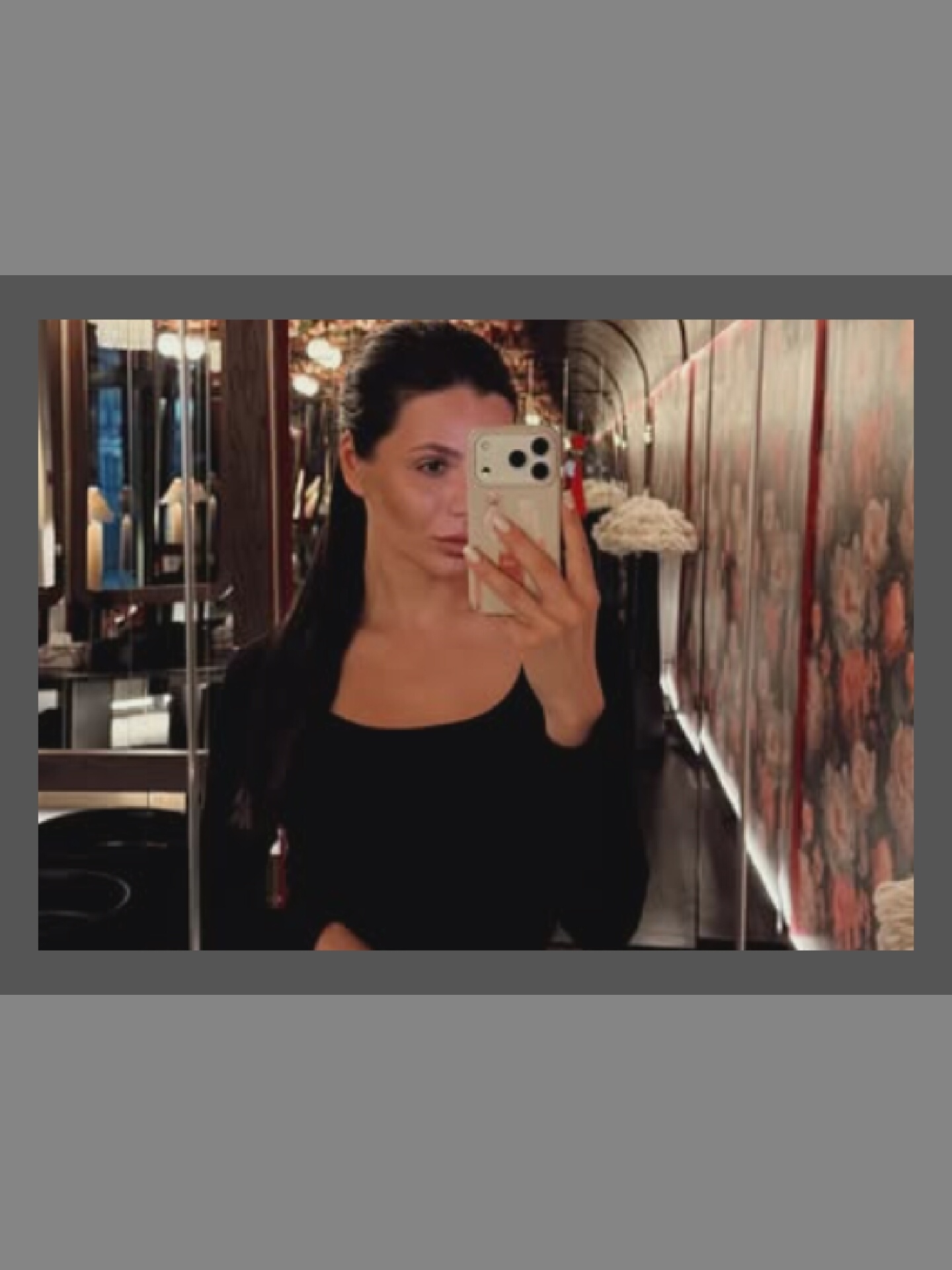 A woman is taking a selfie in a dimly lit, narrow hallway. She is wearing a black long-sleeved shirt and has her hair pulled back in a ponytail. She is holding a smartphone in her right hand, which is reflecting in a mirror behind her. The hallway has floral wallpaper on the right side and mirrors on the left side, with various objects reflected in them. The floor appears to be dark and reflective.