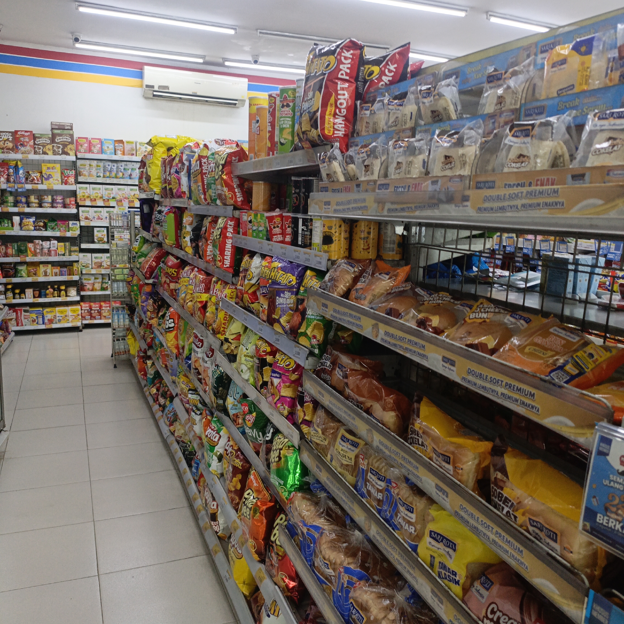 The image depicts the interior of a convenience store, likely a minimarket, in Salatiga, Indonesia. The perspective is from the aisle, looking down a row of shelves stocked with various food products. The shelves are made of metal and display an array of packaged snacks, chips, bread, and other convenience items. The products are brightly colored and feature Indonesian language packaging. On the left side of the image, shelves are filled with cereal boxes and smaller packaged goods. The aisle floor is tiled with light-colored squares. Overhead, fluorescent lighting illuminates the scene, and an air conditioning unit is visible on the wall. The overall impression is of a well-stocked and organized retail environment.