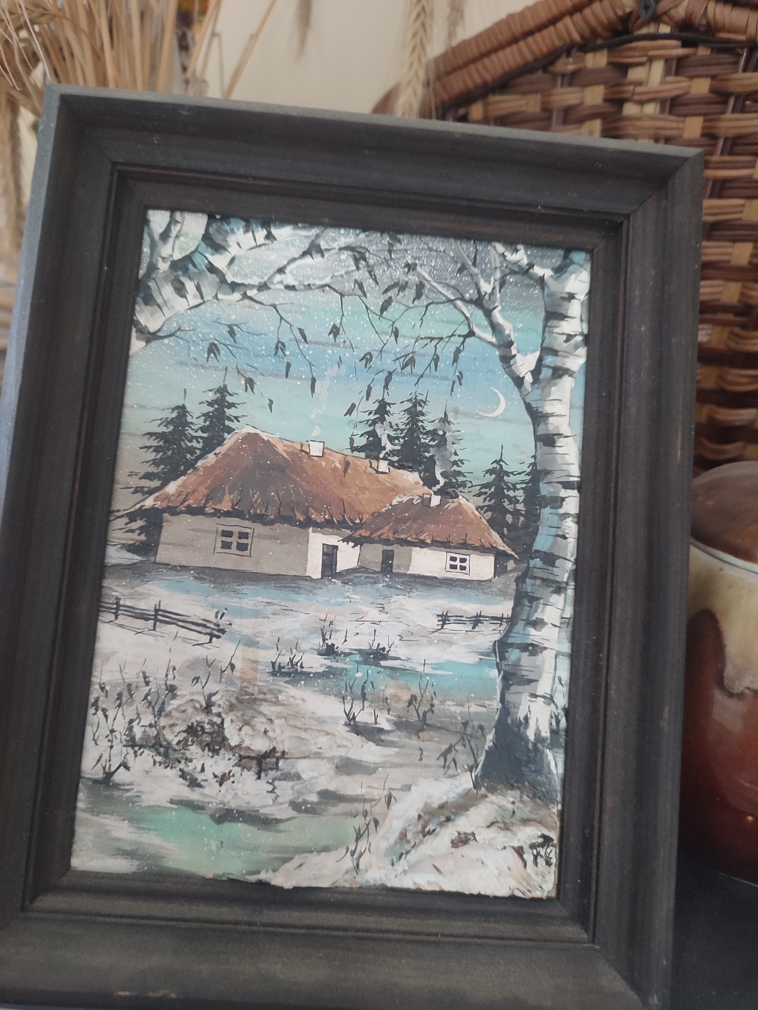This is a framed painting depicting a winter scene in Kriukivshchyna, Ukraine. The painting shows two traditional Ukrainian houses with thatched roofs, covered in snow. In the foreground, there is a snowy landscape with bare trees and a wooden fence. A large birch tree with distinctive white and black bark is prominent on the right side of the painting. Evergreen trees are visible behind the houses. The sky is a pale blue with a crescent moon, suggesting it is either dusk or early dawn. The overall mood is peaceful and serene, characteristic of a quiet winter day. There is no visible text in the painting. The painting is housed in a dark, wooden frame.