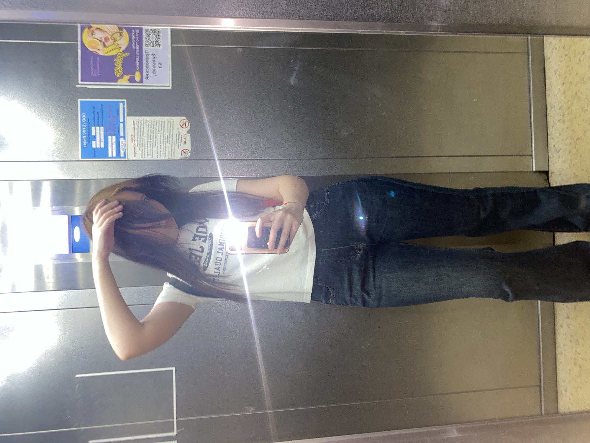 A young woman stands in an elevator and takes a selfie with her smartphone. She wears a white short-sleeved shirt, dark blue jeans, and has long, dark hair. The elevator interior is gray and metallic. There are two advertisements and a sign in Cyrillic on the elevator wall. The floor is light-colored. The image seems to be taken in an elevator located in Unknown, Unknown.