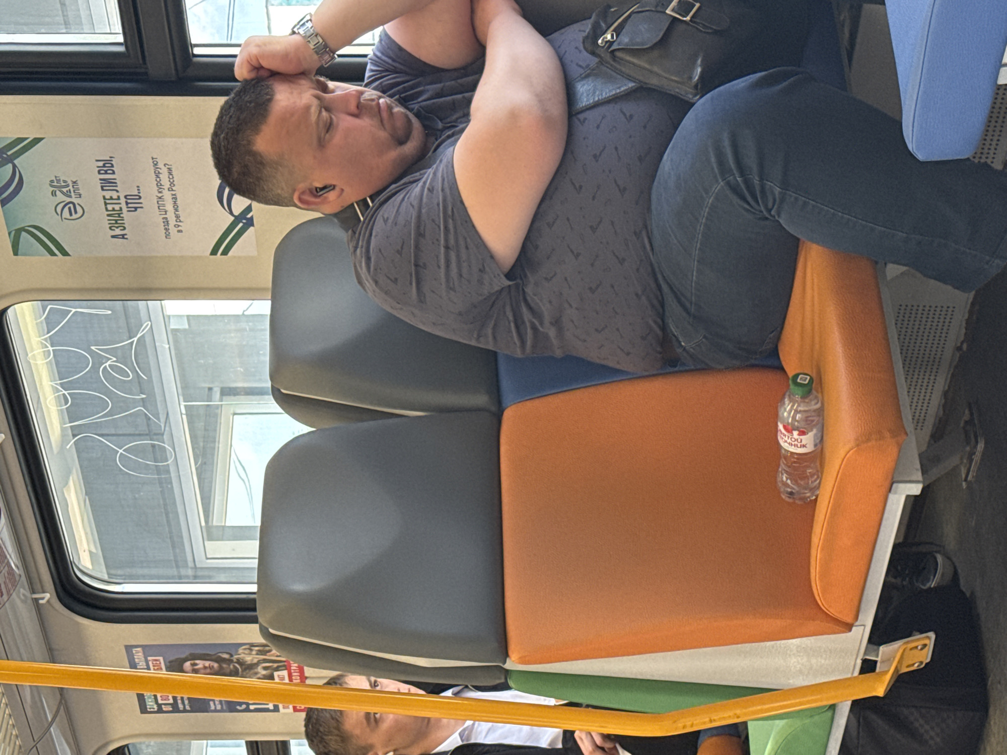 A middle-aged, overweight man with short brown hair, wearing a dark patterned t-shirt and blue jeans, is seated on a train. He has earbuds in and his right hand is on his forehead, suggesting he may be tired or unwell. He is leaning back against the headrest of the seat. Beside him, on the orange seat, is a partially full bottle of water. In front of him, partially visible, is a black bag. The man is seated in a multi-colored seat with orange and gray sections. The train interior features yellow grab handles and windows with some graffiti-like markings on the glass. A poster with Russian text is visible on the wall of the train car, which states that the train company "ЦППК" operates in 9 regions of Russia, hinting at the train's location. This train is likely part of the Central Suburban Passenger Company (ЦППК), which operates in the Moscow Oblast, including the city of Ramenskoye, Russia.