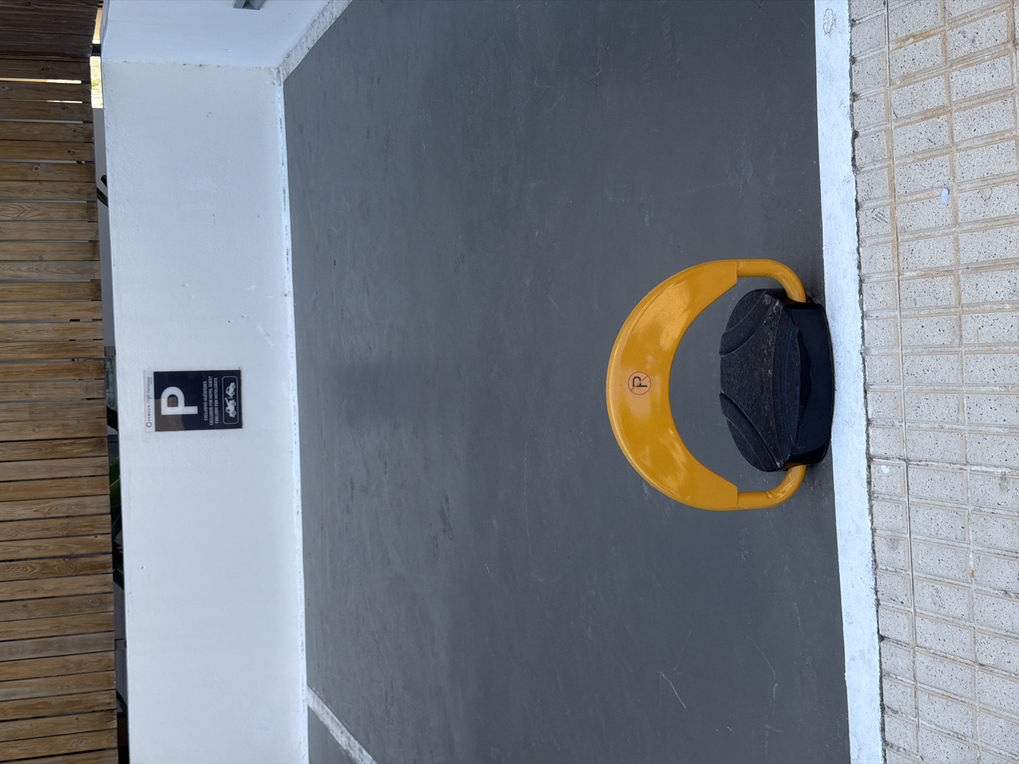 The image shows a parking space in Palmanova, Spain, with a yellow parking barrier in the foreground. The parking barrier is a U-shaped metal arm that can be folded down to block a parking spot. It is black and yellow and has a "P" logo on it. The parking space is paved with asphalt and has a white line marking its boundary. In the background, there is a white wall with a parking sign that reads "P EXCLUSIVO HOTELES GUEST" in English. The wall is topped with a wooden fence. The overall scene suggests a hotel parking area.