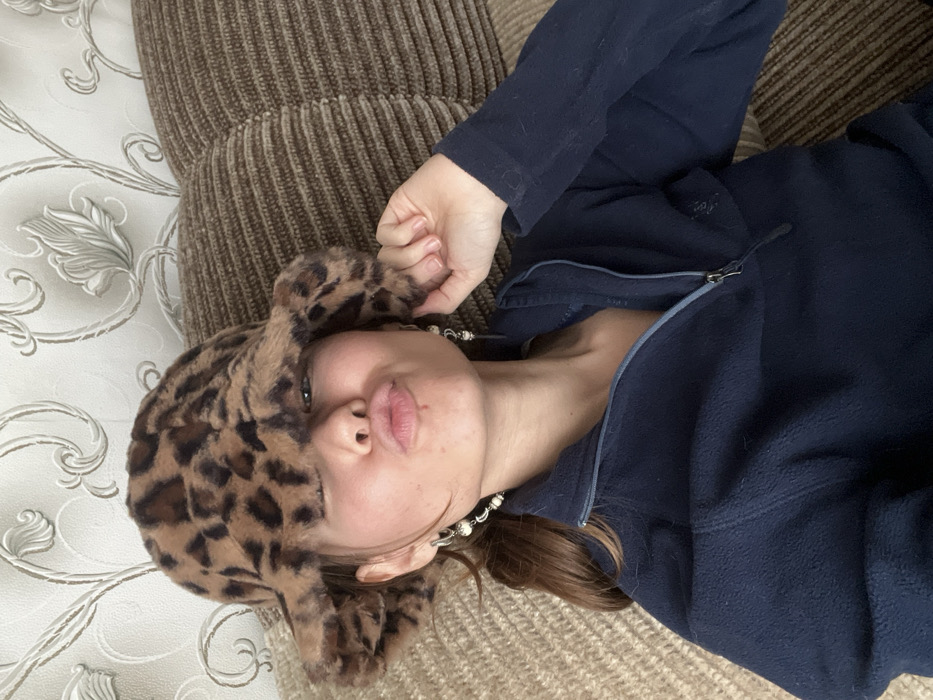 A person lies on a textured beige surface and a brown striped cushion. The person wears a blue zip-up jacket and a leopard-print hat with cat ears. They are tilted towards the camera with pursed lips. A pearl-like necklace adorns their neck. A section of a patterned surface is visible in the upper left corner. The overall composition suggests a casual, cozy setting.
