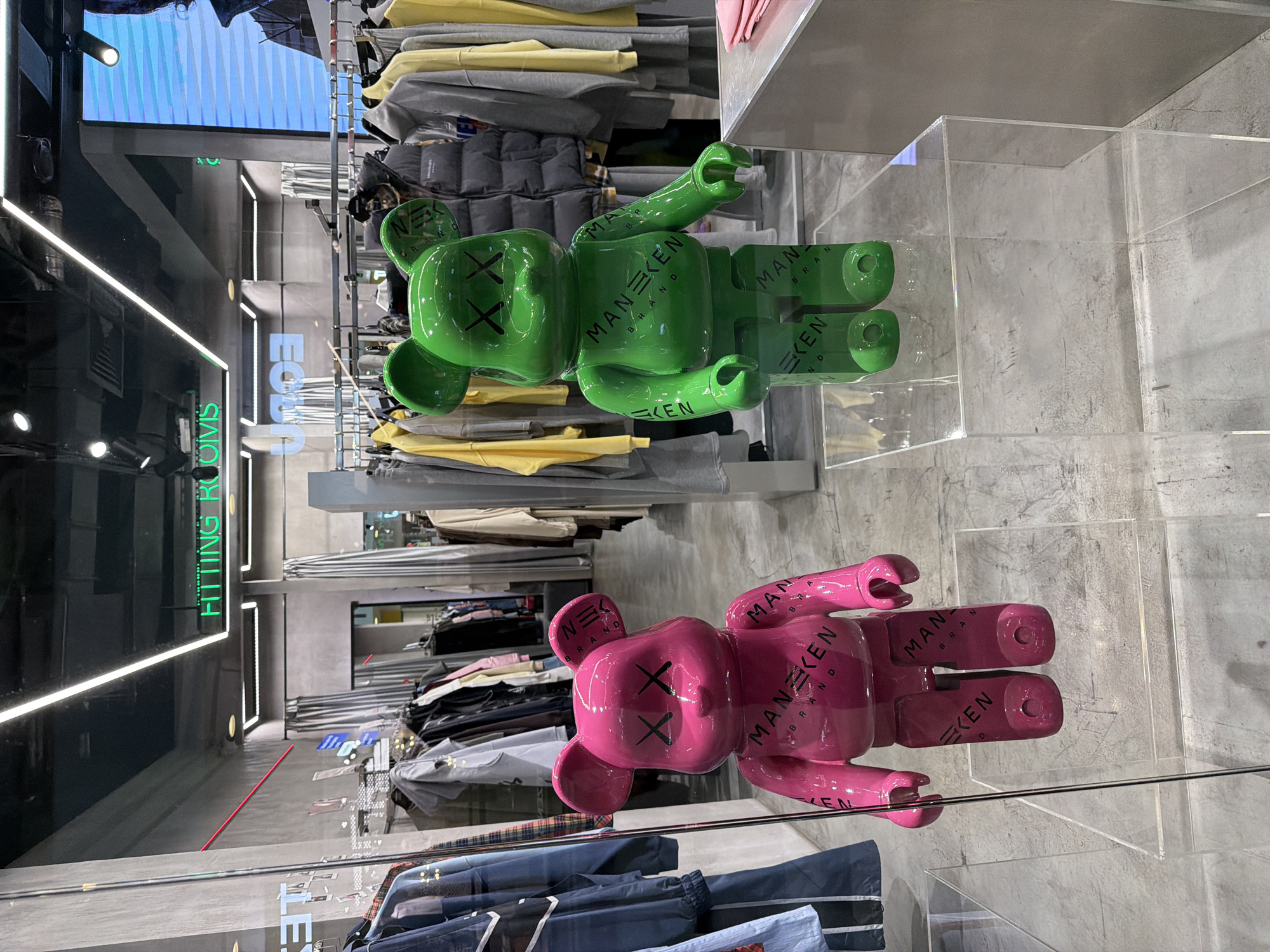 This is an indoor photograph taken inside a retail clothing store. There are clothes on display. In the foreground, there are two bright-colored bear-shaped figures with crossed-out eyes, positioned on transparent platforms. The upper bear is green, and the lower one is pink. Both bear figures have the words "MANEKEN BRAND" printed on them. In the background, the store's interior is visible, featuring clothing racks filled with garments. "FITTING ROOMS" is indicated by bright green lettering on the left. The location of the store is likely in Saint Petersburg, Russia.