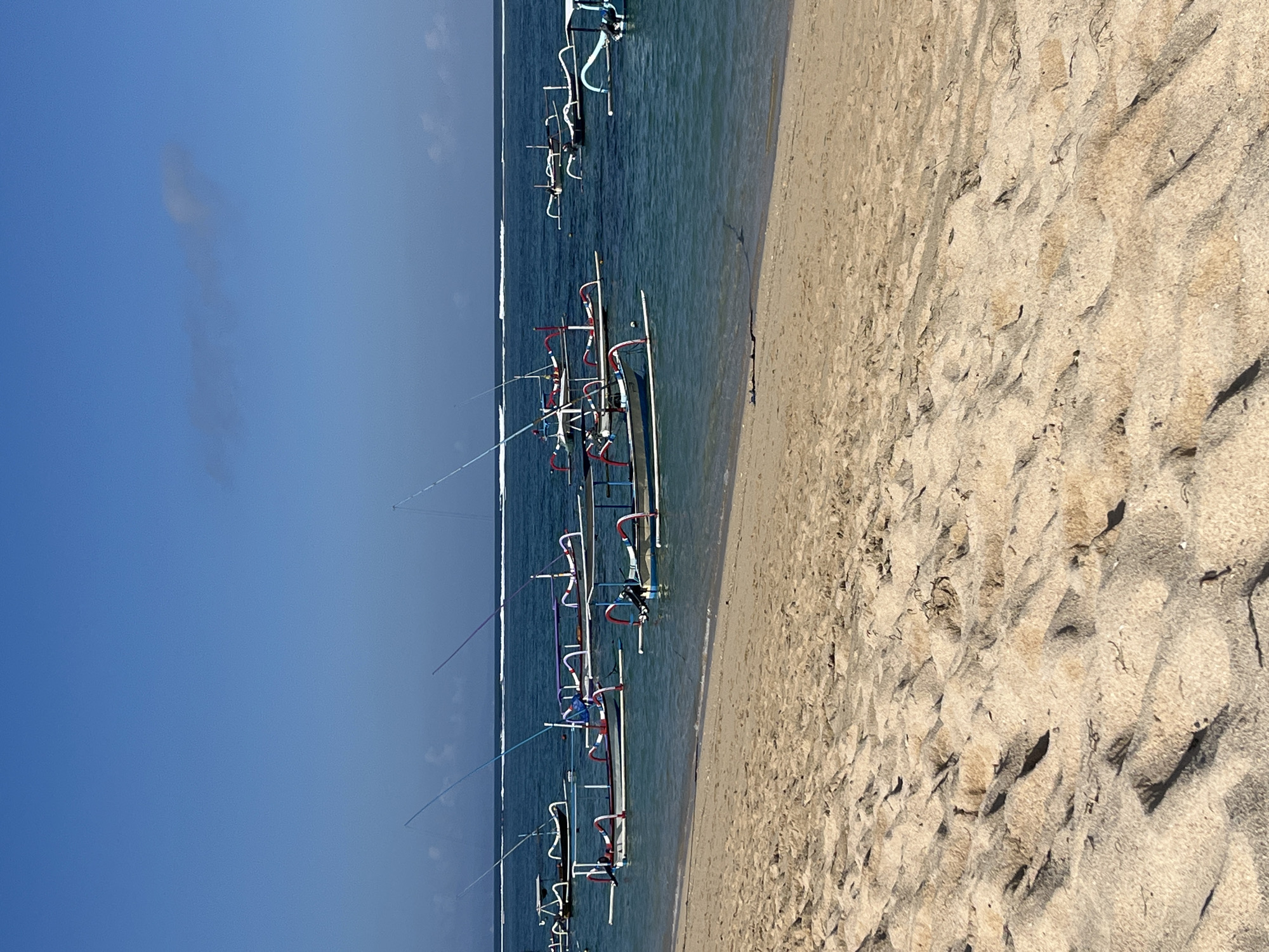 The image depicts a beach scene in Kuta Selatan, Indonesia. The foreground is dominated by a sandy beach with gentle undulations and scattered debris. The middle ground features the calm, turquoise ocean with several traditional outrigger boats anchored offshore. These boats are white with colorful accents, and some have poles extending upwards. The horizon shows a thin white line of breaking waves, separating the sea from a clear blue sky with a faint wisp of a cloud. The lighting suggests a bright, sunny day.