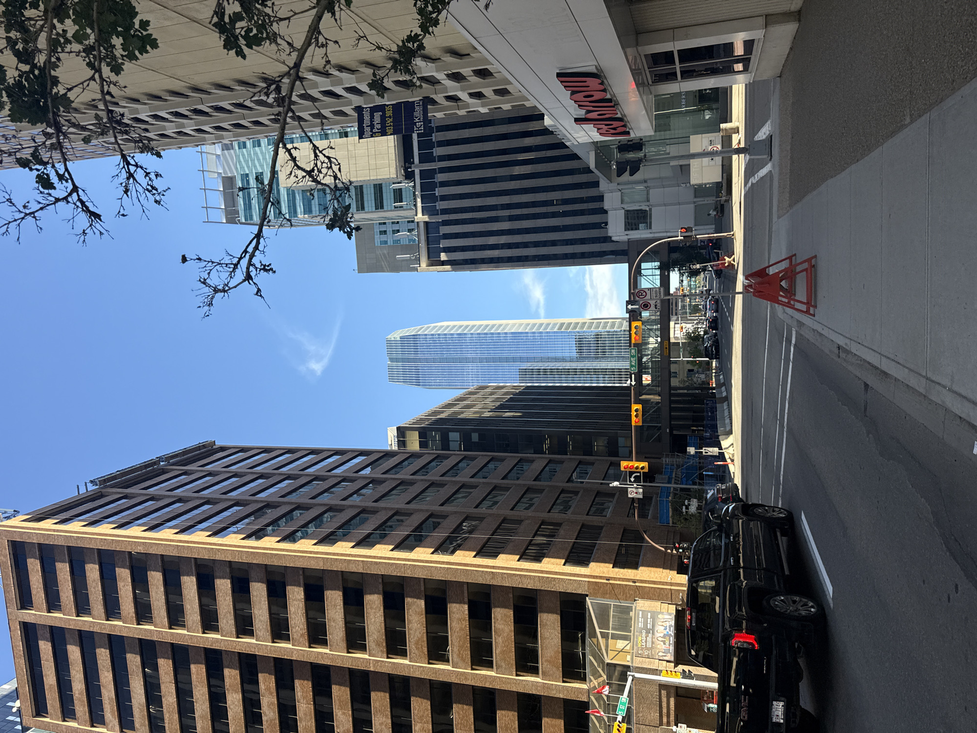 The image depicts a street scene in downtown Calgary, Canada, with a view looking down a city street lined with tall buildings. On the left, a brown brick building with many windows dominates the foreground. To the right of this, a modern skyscraper with a distinctive curved glass facade stands out in the background. Closer to the right side of the image, another building features dark vertical stripes and a sign reading "red arrow".
The street is paved and appears to be a multi-lane road with traffic lights and road signs visible. A black GMC pickup truck is driving in the foreground, facing away from the camera. Other vehicles are present further down the street. Pedestrian sidewalks are visible on both sides of the street, with some orange construction barriers on the right side. The sky is clear and blue with some wispy clouds. Trees with sparse branches are visible in the upper right portion of the image, suggesting it might be a season with less foliage. The overall impression is a busy urban environment on a sunny day.