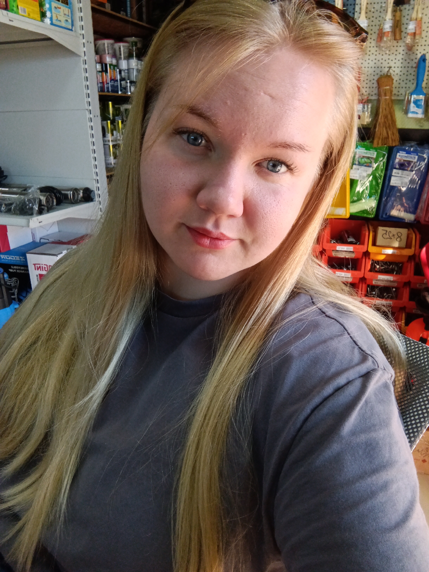 A young woman with long, blonde hair is the primary subject of this close-up, indoor photograph. She has fair skin, blue eyes, and is wearing a gray t-shirt. The woman is looking directly at the camera with a slight smile.
Behind her, the background appears to be a workshop or storage area. A white metal shelving unit is visible to her left, stocked with various items including boxes and cans. To her right, there is a display of hardware supplies, including colorful plastic storage bins filled with small parts, as well as packaged goods and hanging brushes.
There is no information to confirm that the location is in the city of Фоки, Russia.