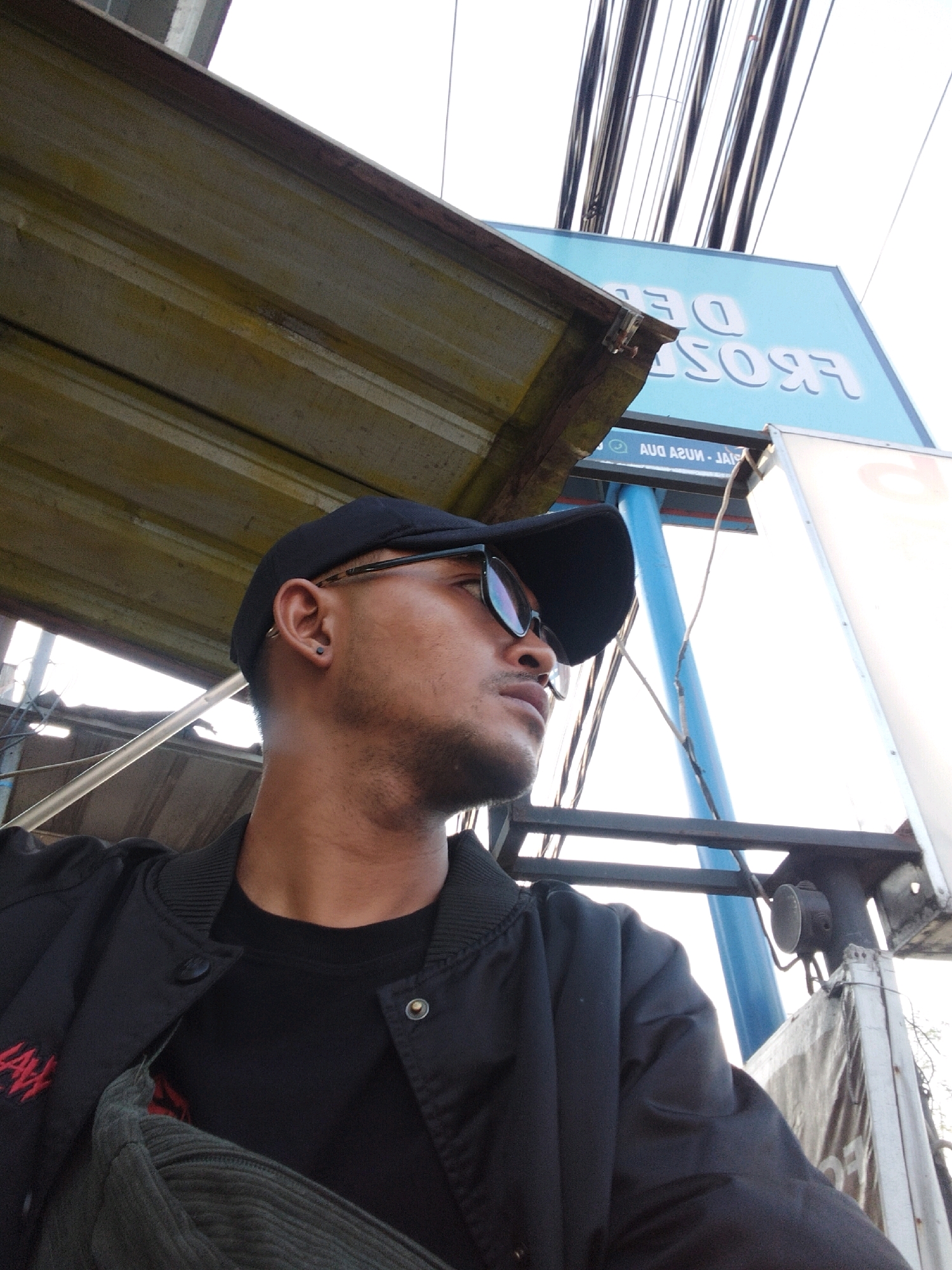 In a medium shot, a young man wearing a black baseball cap, dark sunglasses, a black jacket over a black t-shirt, and a grey corduroy bag across his chest is seen from the chest up. He is positioned on the right side of the frame, looking to the left. The man has a light stubble and is wearing a small black stud earring in his left ear. Behind him, a portion of a blue sign with white lettering is visible, reading "DE PROSE", and below that, in smaller text, "DIAL-NUSA-JAIS". The sign is partially obscured by a corrugated metal awning overhead. The background consists of an overcast sky with numerous electrical wires crisscrossing the scene. The overall environment suggests an outdoor urban setting, possibly a street or market area in Kuta Selatan, Indonesia.