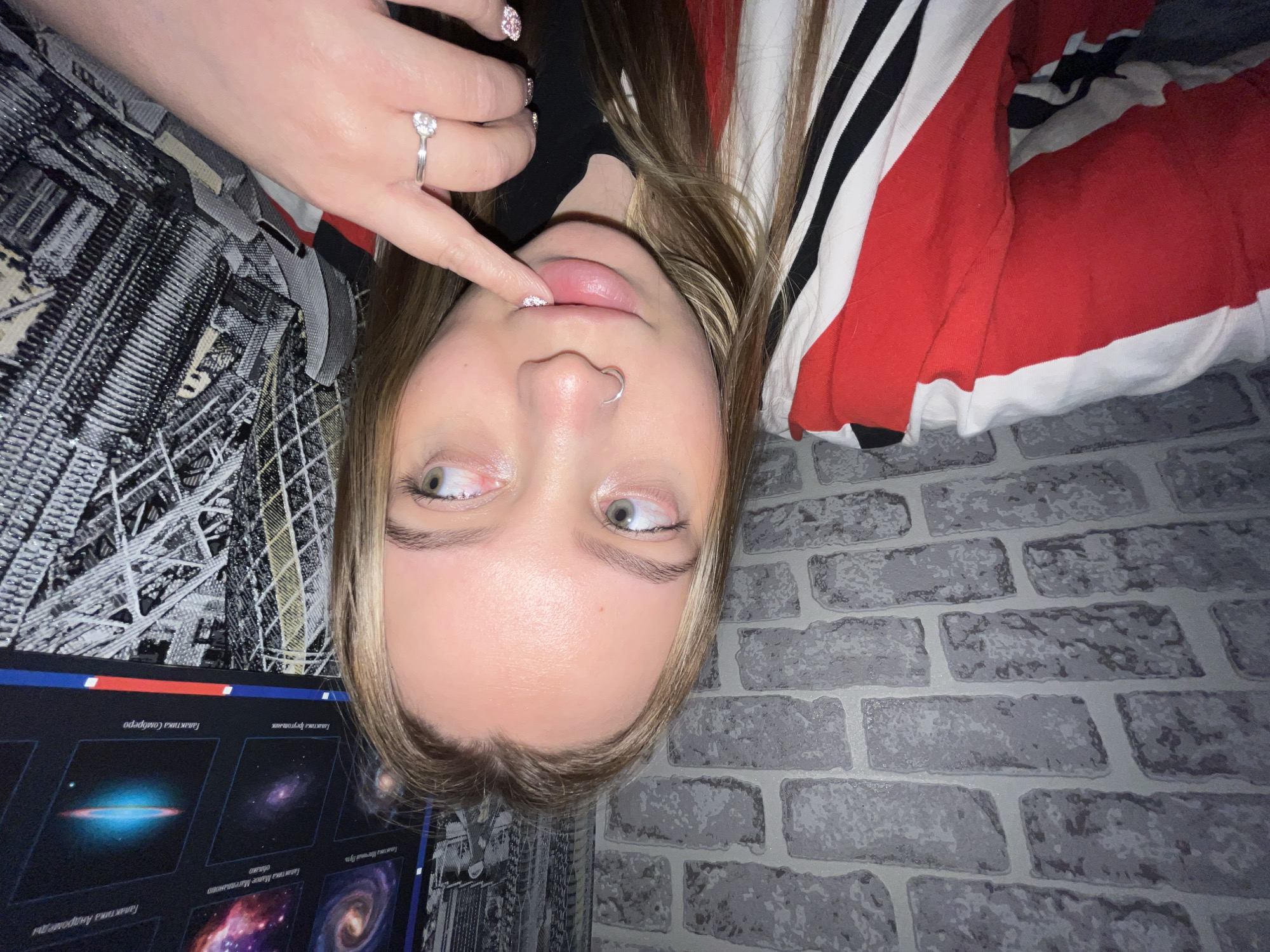 A young woman's face, inverted, is centered in the frame. Her left index finger, adorned with glittery pink nail polish and a ring, is touching her upper lip. She has a nose piercing and fair skin. She is wearing a black top beneath a red, white, and black striped garment. The background includes a brick-patterned wall and a patterned fabric. A poster with images of galaxies is visible. The location in the city of Novopokrovskaya, Russia, cannot be determined from the image.