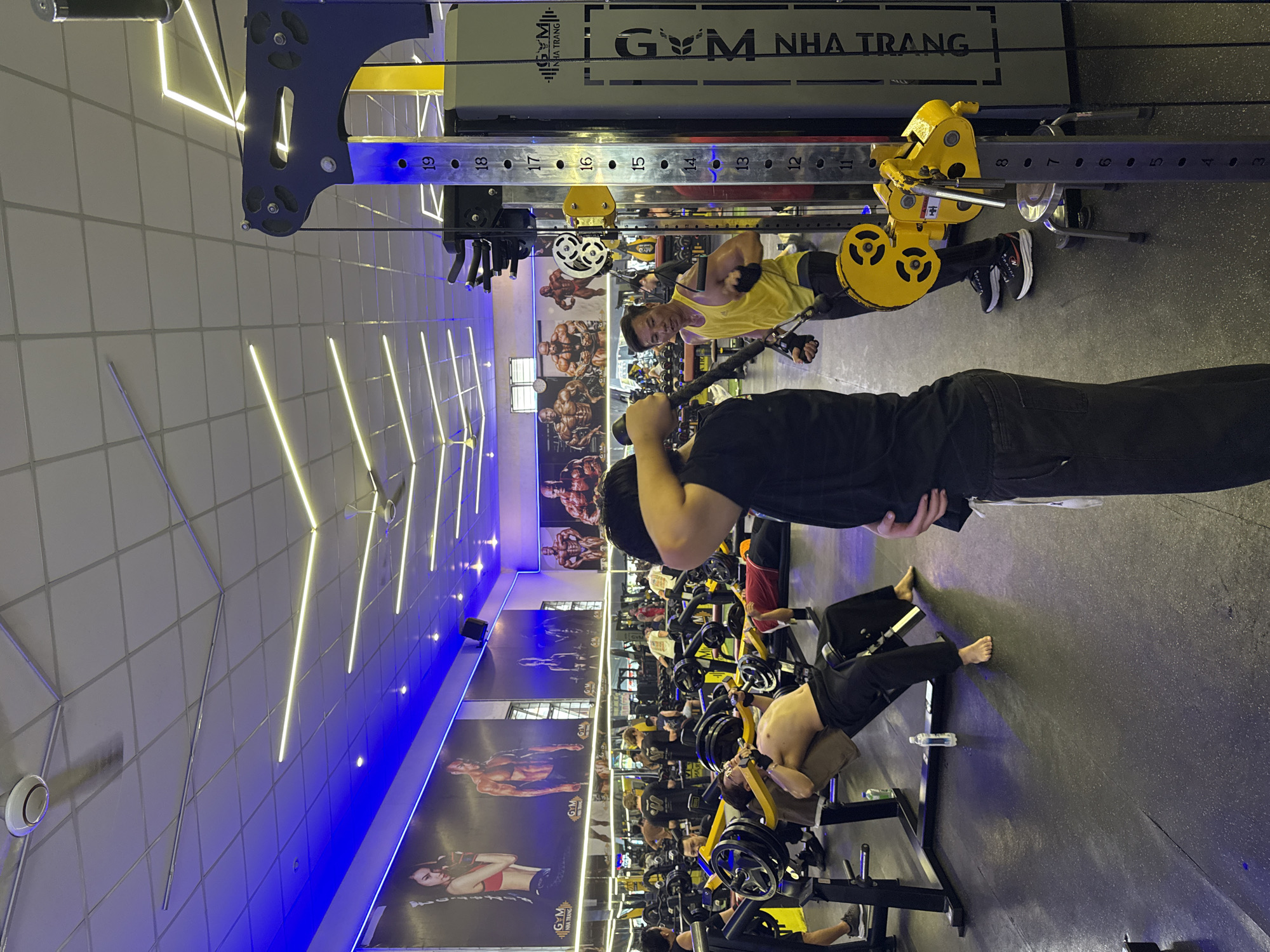 The image depicts the interior of a brightly lit gym with a modern design. Several individuals are engaged in various workout routines. In the foreground, a man wearing a yellow tank top and black workout pants is performing a cable exercise, gripping a handle with both hands. Behind him, another person in black is lying on their back, also engaged in a workout with a cable machine. Further back, multiple individuals can be seen on different exercise machines, including weightlifting equipment. The gym is decorated with large murals of muscular figures on the walls, and neon lights, predominantly blue and yellow, illuminate the space. The text "GYM NHA TRANG" is visible on a piece of equipment in the upper part of the image. The overall atmosphere suggests an active and energetic gym environment. The time of day is not explicitly indicated, but the lighting suggests it could be evening or a well-lit indoor space regardless of the external time.