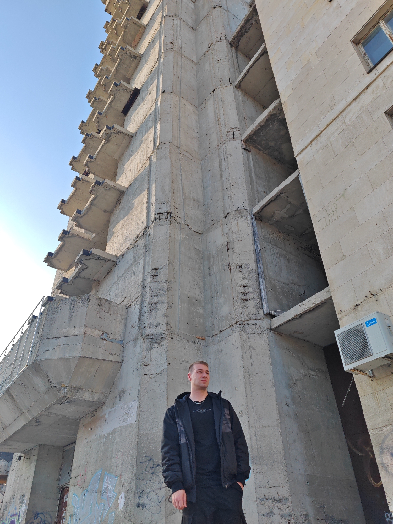 A light-skinned adult male stands on the ground, positioned against the base of a large, brutalist-style concrete building. He is wearing a black hooded jacket over a black t-shirt with white "MINDSTORM" text, and black pants, with his hands in his pockets and his gaze directed towards the viewer's left. The structure behind him is the unfinished or brutalist-style concrete section of the Youth House (Младежки дом) complex in Shumen, Bulgaria. The building exhibits an extensive facade on the left composed of numerous exposed, staggered, cantilevered concrete balconies or platforms, showing raw concrete, visible rebar, and signs of weathering. The central and right sections of the building consist of rough concrete with uncompleted construction elements. A portion of the building on the right features a finished facade of light-colored rectangular masonry blocks, containing a closed window with a blue tint and a white "Midea" brand outdoor air conditioning unit mounted below it. The lower concrete walls of the structure are covered in various spray-painted graffiti. The sky above is clear blue, indicating daylight conditions.