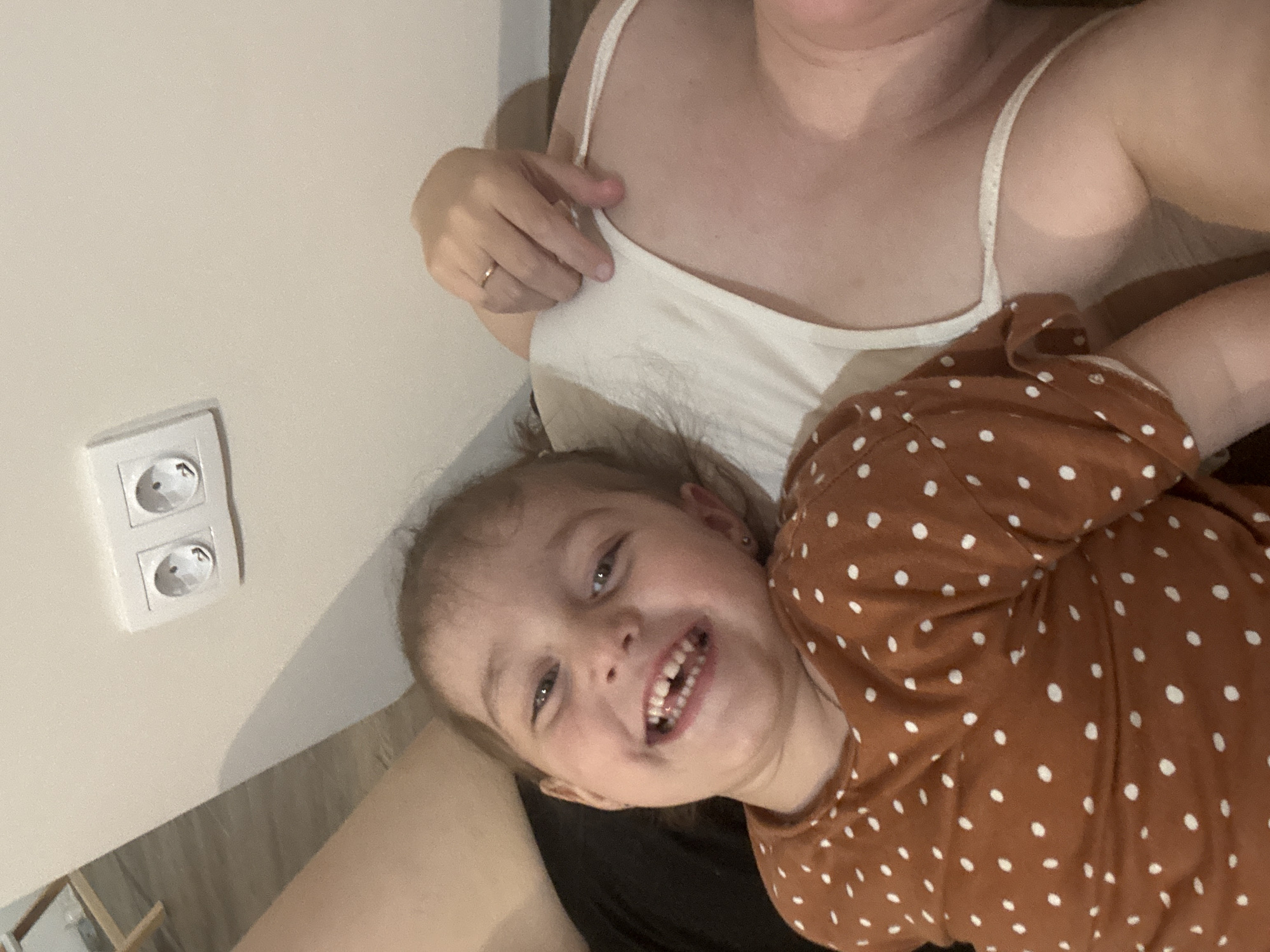 A close-up shot features a fair-skinned child with light brown hair and brown eyes, smiling broadly with visible teeth. The child is wearing a brown, long-sleeved shirt with white polka dots. The child is situated on the lap of an adult with fair skin who is wearing a white tank top. The background includes a light beige wall with a white electrical outlet. The image was taken indoors. There is no data regarding the location within the city of Chaikovsky, Russia.