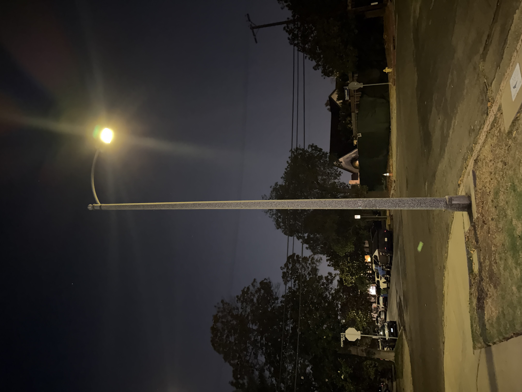 The image depicts a night scene on a street in an unknown city. A tall, gray streetlight dominates the center of the frame, its bright yellow-green light illuminating the surrounding area and creating a lens flare. The sky is dark blue with a few faint stars visible. Trees with dark green foliage flank the street, casting shadows. A few cars are parked on the side of the road and on the street. In the background, houses with illuminated windows and a tall utility pole with power lines can be seen. The street itself is paved and appears to be an intersection. A street sign reading "PICKFORD ST" is visible on the left side of the image. A fire hydrant and a traffic cone are also present in the scene. The overall impression is that of a quiet residential street at night.