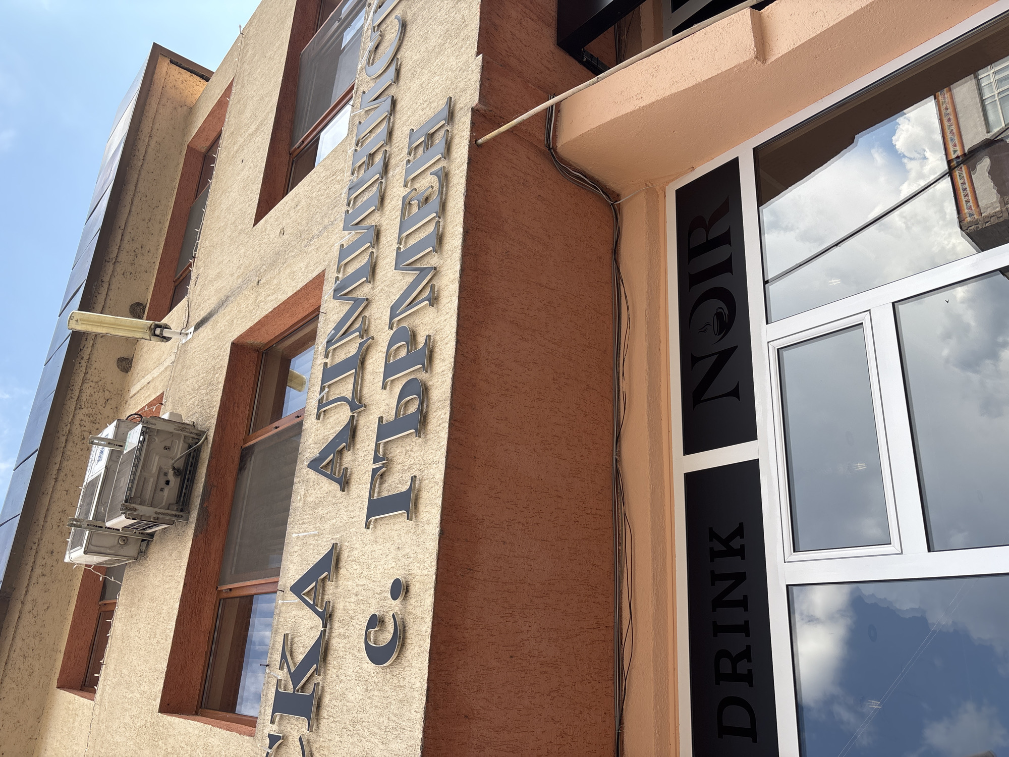 The image shows a building in Garmen, Bulgaria. The building has a beige facade with brown window frames. The name "С. АУМИС ГРМЕН" is vertically written in Cyrillic letters on the wall. An air conditioning unit is mounted on the wall. Next to the building, a sign on a window reads "DRINK NOIR". The sky is visible in the background. The reflection of a nearby building is visible in the glass of the "DRINK NOIR" window. Some electrical wires are running along the side of the building.