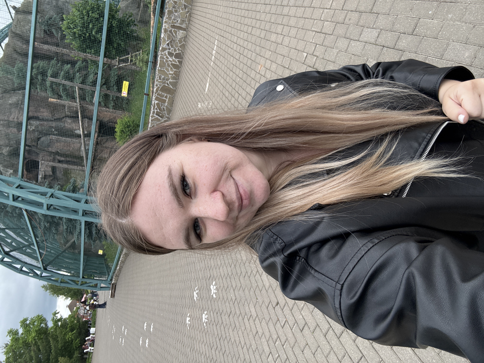 A selfie-style photograph captures a young adult female in the foreground, centered and slightly to the right, against an outdoor park or zoo environment. The woman has light brown hair with blonde highlights, parted on the left side, and fair skin with visible freckles. She is smiling faintly towards the camera, revealing light-colored eyes. She wears a black faux leather jacket with a zippered front and metallic buttons. Her right hand is visible in the lower right corner, clenched lightly, with white nail polish on her fingernails.
The background depicts a paved pedestrian walkway made of grey interlocking pavers. On the left side of the walkway, a series of white animal paw prints are painted on the ground, leading into the distance. Further back on the left, several indistinct individuals are visible, some seated on benches, amidst green trees and distant buildings under an overcast sky.
On the right side of the image, a large animal enclosure is prominent. It features a robust, green metal arching structure with mesh fencing, housing natural elements like large rock formations, numerous evergreen trees, and fallen logs. A wooden ladder is visible against one of the rock structures within the enclosure. A small yellow warning sign with a black exclamation mark in a triangle and Russian text (partially legible as "ОСТОРОЖНО" - Caution) is affixed to a lower metal bar of the enclosure. The base of the enclosure is lined with a decorative stone wall composed of various sized and colored rocks. This scene is consistent with the Izhevsk Zoo in Izhevsk, Russia, given the animal enclosure, paw prints, and Russian signage.