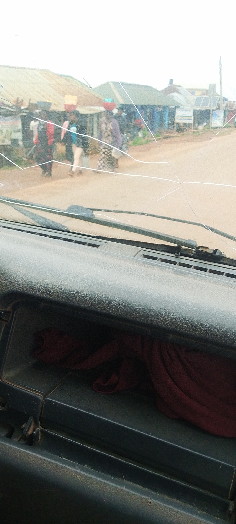 This media file captures a view from inside a vehicle, looking through a cracked windshield at a street scene in Reifild-Ist, Nigeria. Several people are visible walking along a dirt road in front of various market stalls or small shops. The people appear to be carrying baskets or loads on their heads, suggesting they are engaged in commerce or transport. The buildings in the background have tin roofs and appear to be simple structures. The overall impression is that of a bustling marketplace or a busy street in a rural or semi-urban African setting. The weather appears to be overcast or hazy, as the sky is bright but lacks distinct clouds or direct sunlight. The time of day is not clearly discernible, but the activity suggests it is likely daytime, possibly mid-morning or afternoon. The cracked windshield is a prominent detail, indicating the vehicle may be older or has experienced some damage. There are no clear signs or text legible in the image that would provide further specific location details or context. The scene evokes a sense of everyday life and activity in the community.