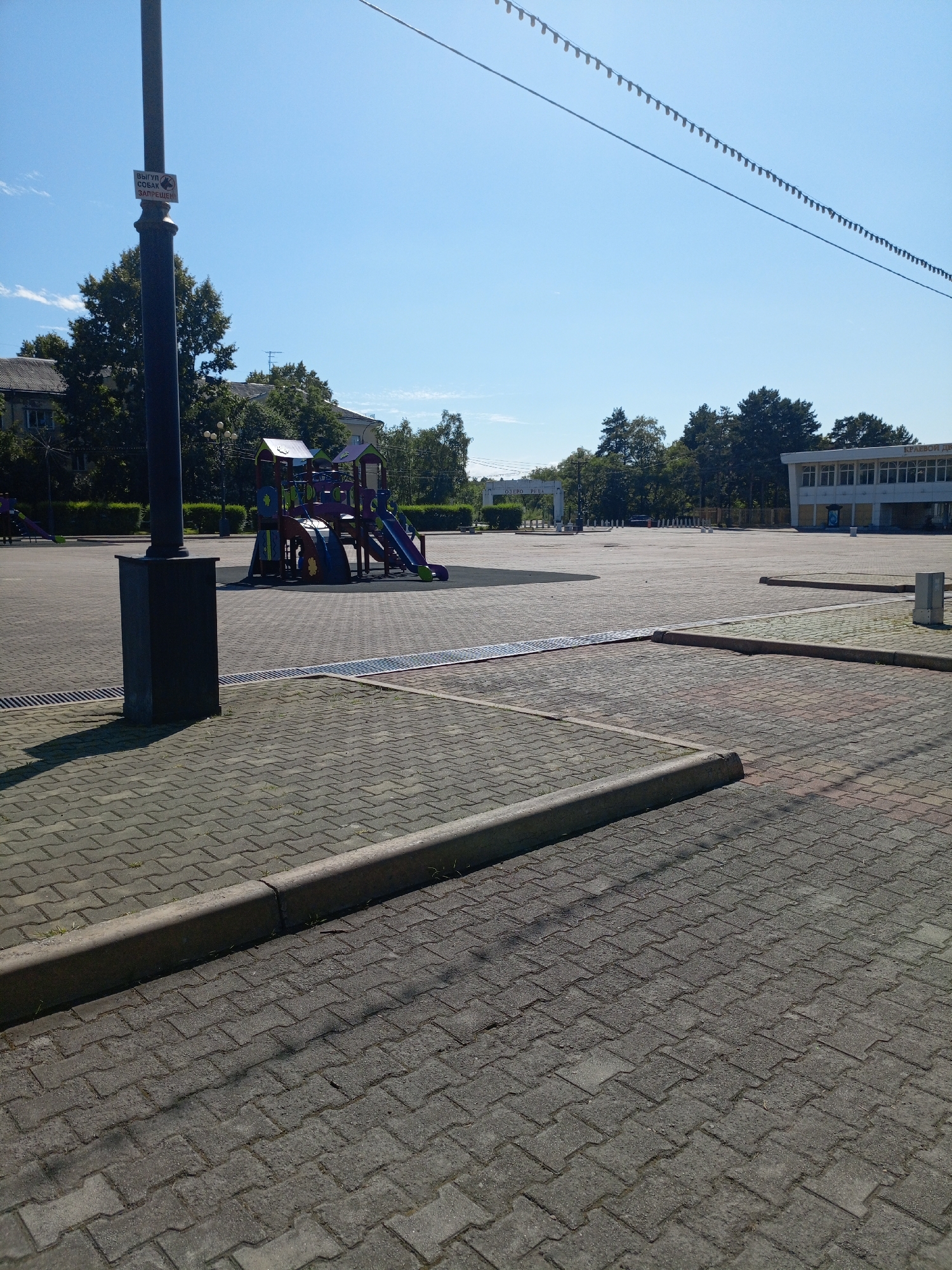 The image depicts an outdoor paved area, likely a public square or plaza, on a clear day. The foreground is dominated by textured grey paving stones, with a raised concrete curb running along the bottom edge of the frame. A dark grey rectangular structure, possibly a plinth or base, is visible on the left, casting a shadow. A tall, dark pole, likely a lamppost, stands behind this structure and extends upwards out of the frame, with a small white sign attached to it bearing Cyrillic text and an image of a dog with a red line through it, indicating that dogs are prohibited.
In the middle ground, a colorful playground structure with slides, ramps, and climbing elements is set up on a dark, soft surface. Beyond the playground, the paved area continues, with some trees and buildings visible in the background. The buildings appear to be modern, with glass windows. A string of decorative flags or lights is strung across the upper portion of the image against a bright blue sky. The scene is quiet and appears to be unoccupied by people.