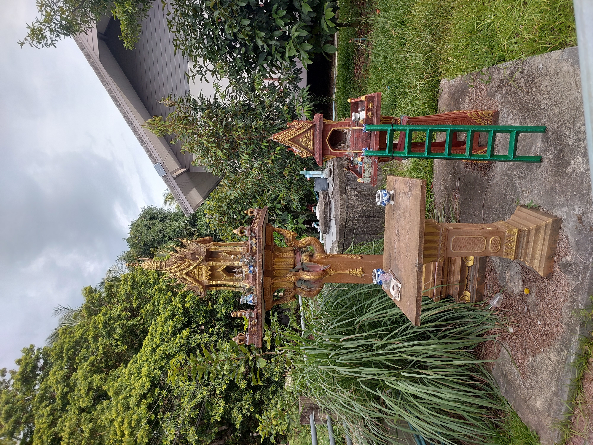 In Baan Mae Nam, Thailand, two miniature spirit houses stand on a concrete platform. The larger, golden spirit house is intricately decorated with Naga serpents and ornate roofs, resting on a tall, cylindrical pedestal. To its right, a smaller, reddish-brown spirit house is adorned with a golden, tiered roof. A bright green ladder leans against this smaller structure.
The scene is set outdoors, with lush green trees and grass surrounding the spirit houses. In the background, a modern building with a grey, angled roof is visible. The sky is overcast, suggesting a cloudy day. No people are present. The objects include the spirit houses, the ladder, a small white and blue ceramic cup on a wooden plank, and scattered debris such as dried leaves and a small plastic cup on the ground. The overall impression is one of a tranquil, possibly sacred, outdoor space.